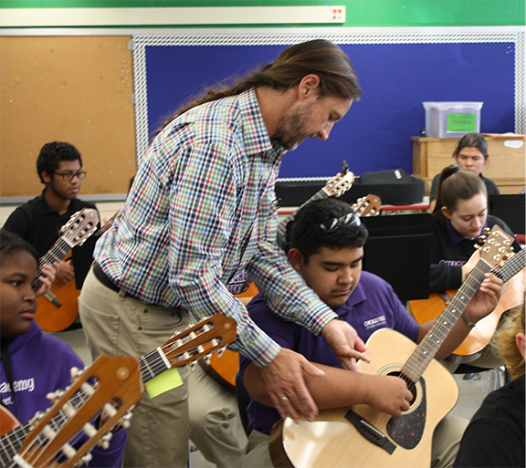 teacher helping student play guitar in a classroom