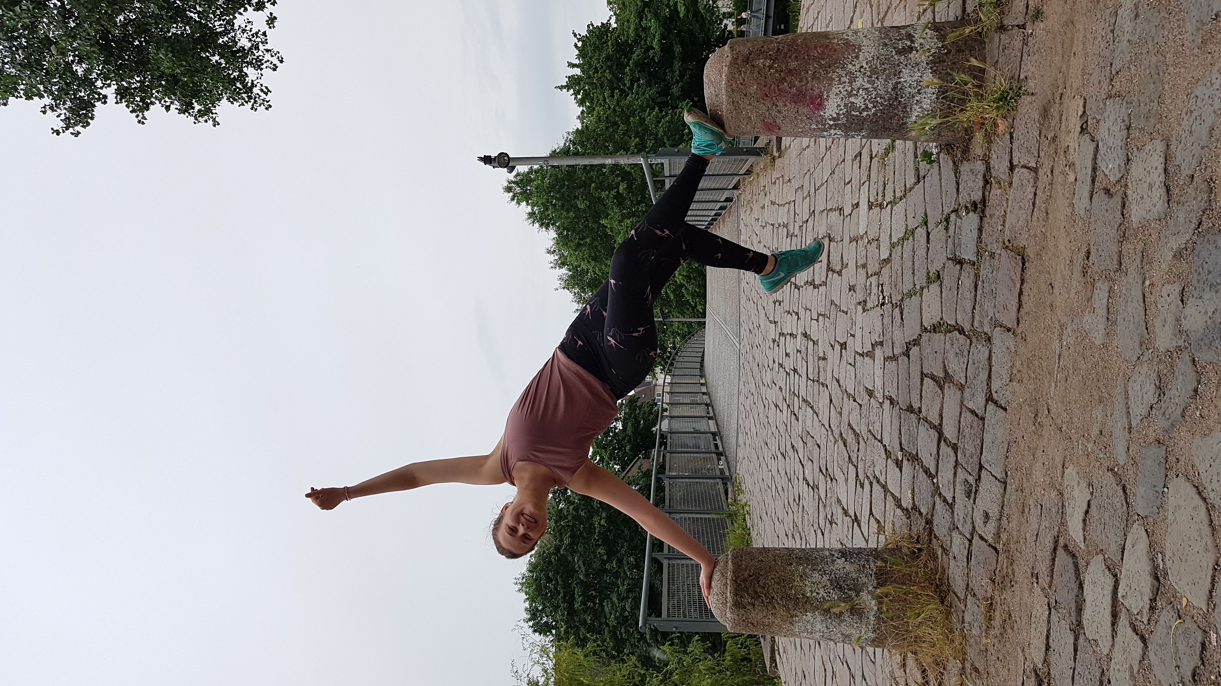 Zwei Poller vor einer Brücke, Frederike stützt sich mit einer Hand auf dem einen, mit einem Fuß auf dem anderen ab und springt herab.