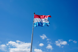 Luxembourgish flag – of red, white and light blue stripes – fluttering in the wind at the top of a flagpole and against a clear blue sky with a few white clouds
