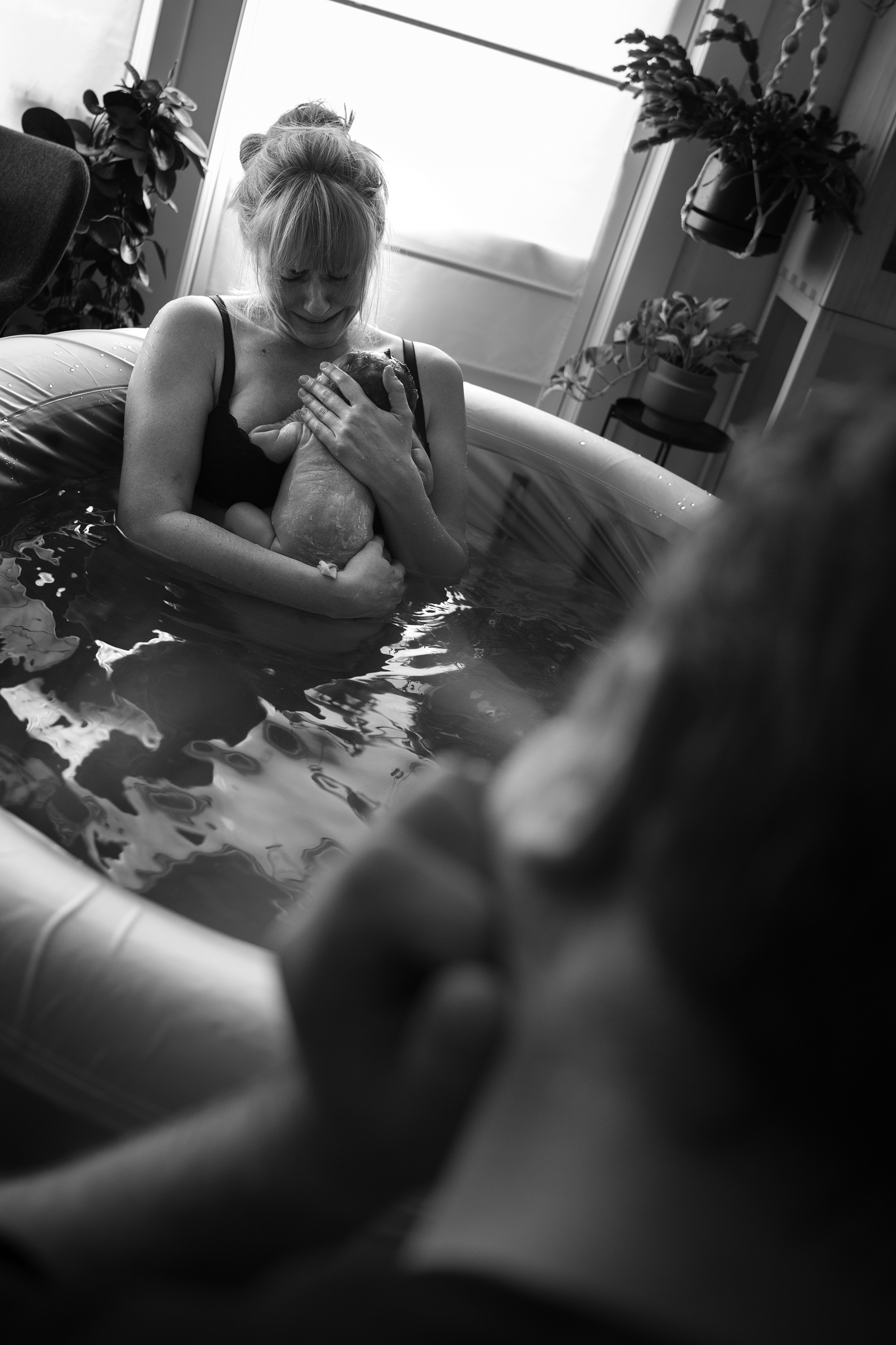 A woman in the birthpool is holding her newborn baby, and feeling emotional. The partner is by the pool, admiring them both