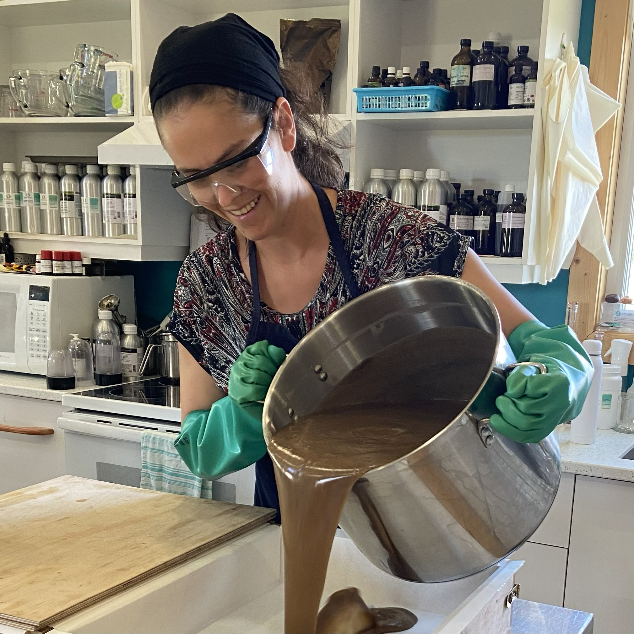 The soapmaker is pouring soap wearing all safery apparel: gloves, goggles and a wide bandana to cover her hair.