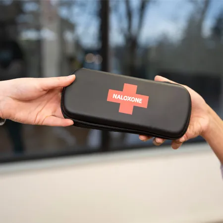 The hands of a person handing a Naloxone kit to another person. 