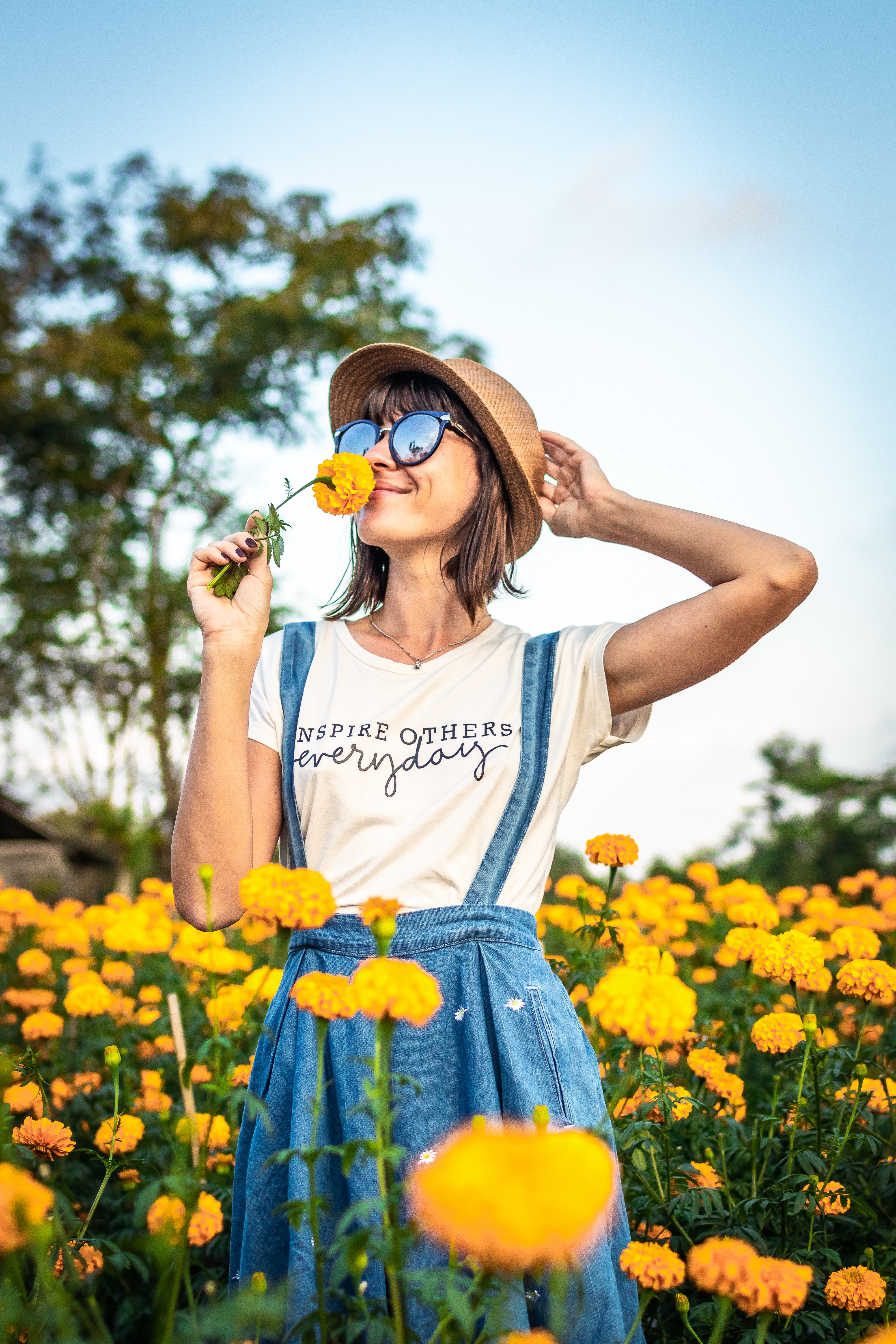 smiling young woman with sunglasses and hat in meadow of yellow flowers smelling flower