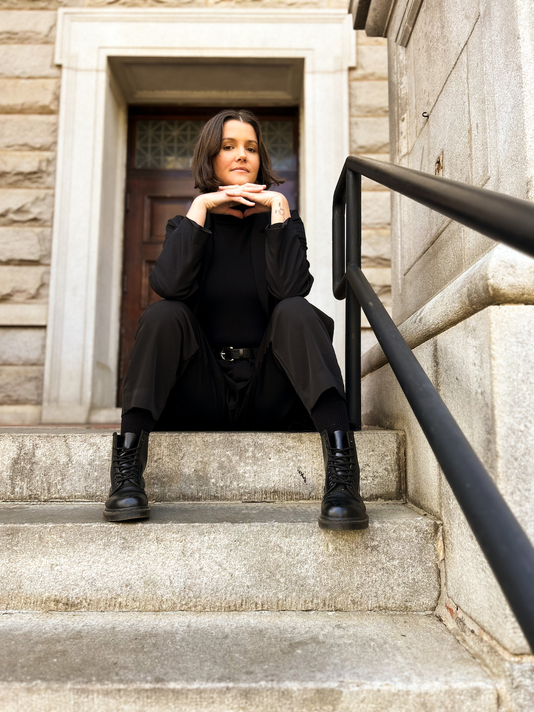 Malerie, a disabled and chronically ill yoga teacher, sits on the top stair with a gentle smile-less expression.