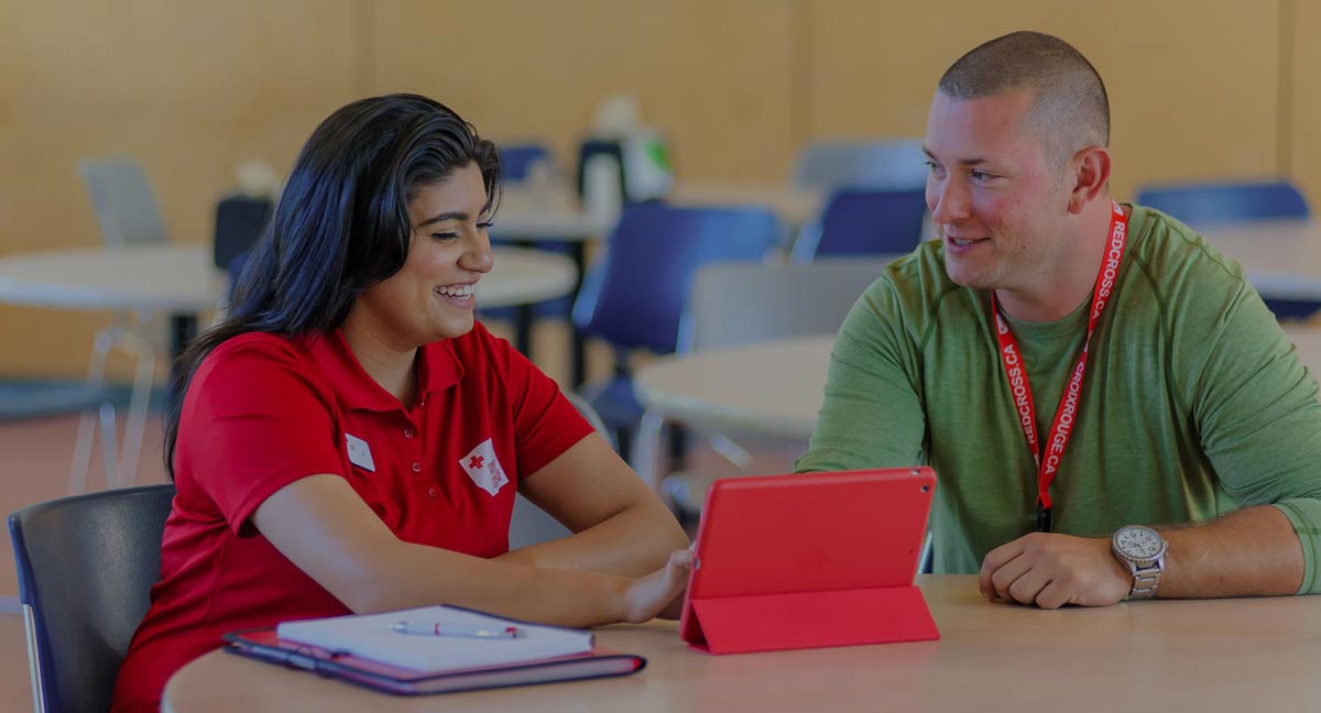  Une personne monitrice de secourisme portant un chandail de la Croix-Rouge assise à une table consulte une tablette en compagnie dune personne apprenante.