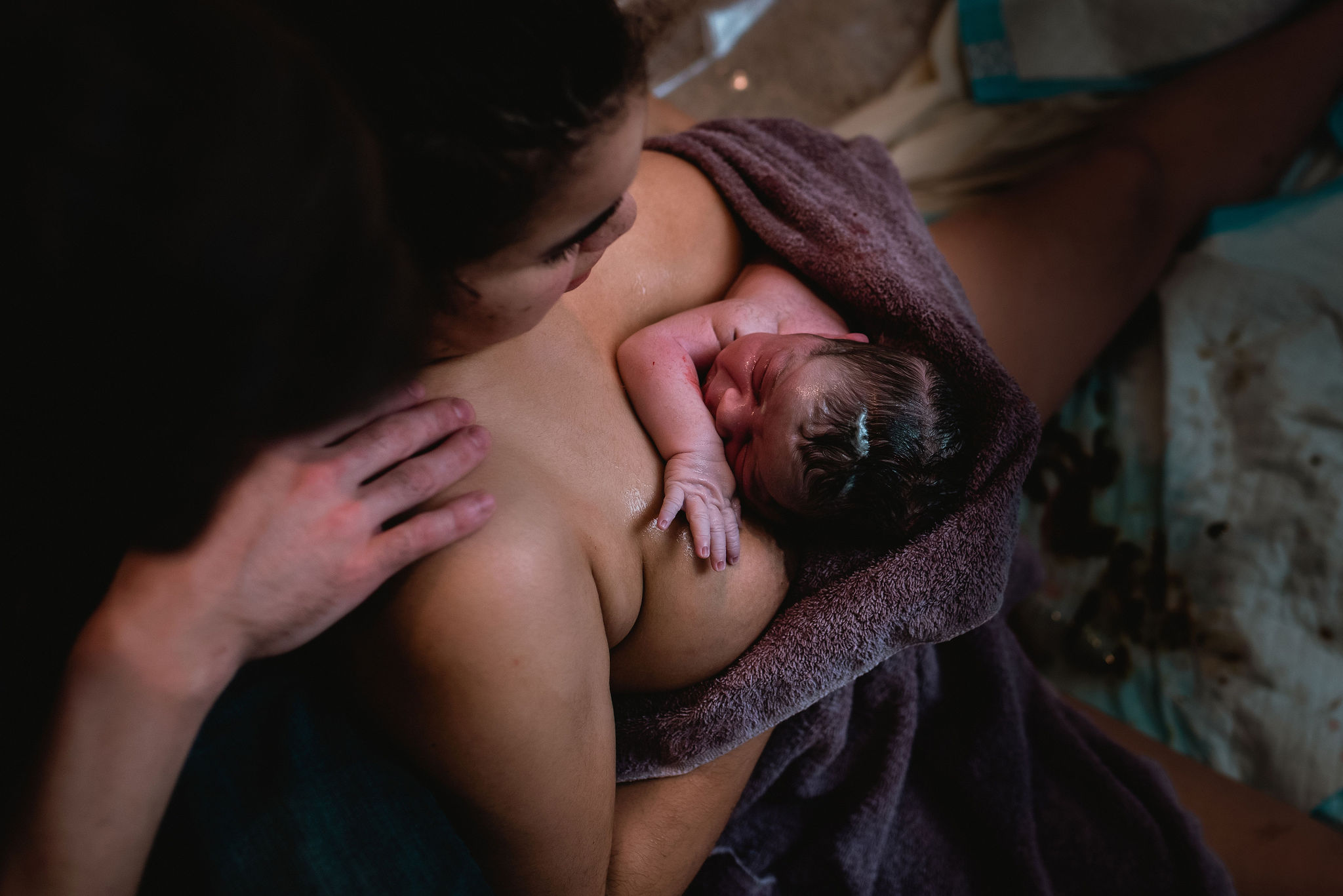 A new parent looks down at their newborn baby on their chest wrapped in a brown towel. There is a hand on the birthing person