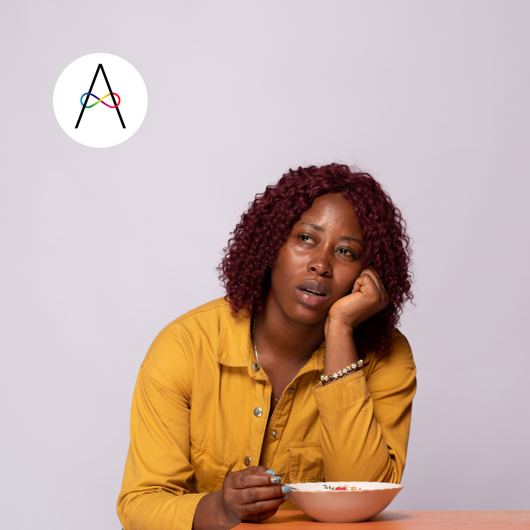 Picture of a black woman sitting at a table with a bowl, she is holding a spoon and gazing into the distance with a vacant expression on her face