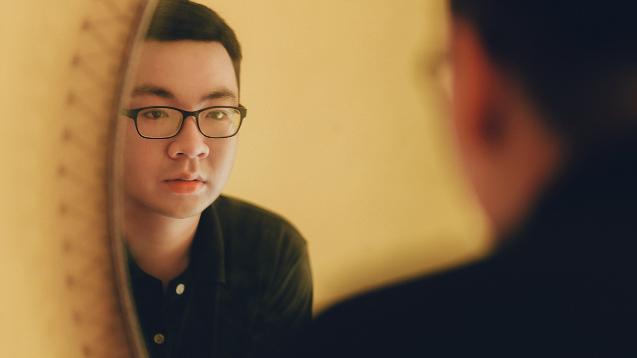Picture of an Asian man wearing glasses looking at his reflection in a mirror