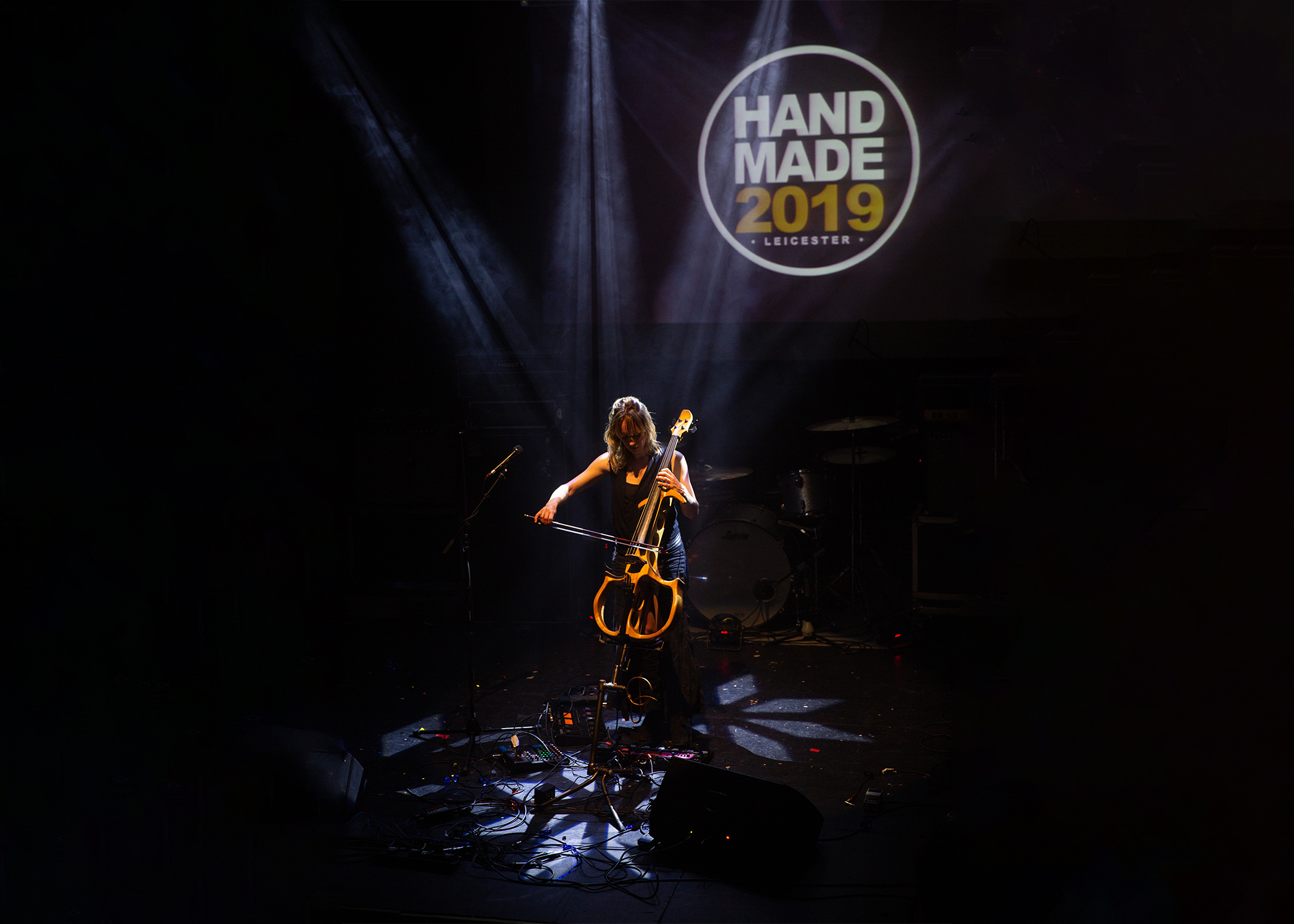 A woman playing an electric cello, lit from above, the light creating patterns on the floor.  A logo in the background reads Handmade 2019.