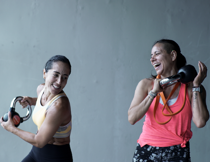 Dos mujeres con ropa deportiva y sonrientes entrenan con kettlebells en la mano.