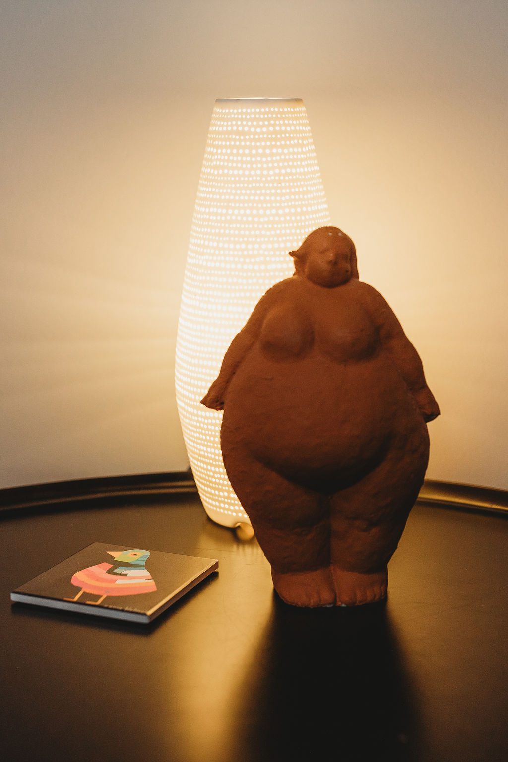 Lamp providing soft light on a black table, coaster amd clay statue of a fat female body 