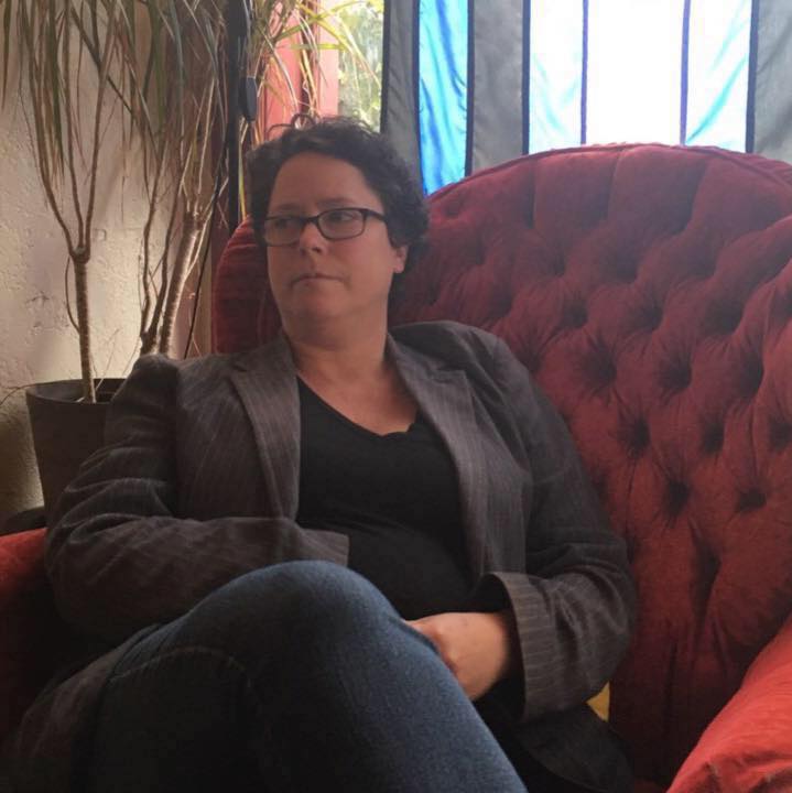 Picture of Miriam Green wearing a grey blazer, black shirt and blue jeans in an overstuffed red chair at Wicked Grounds.