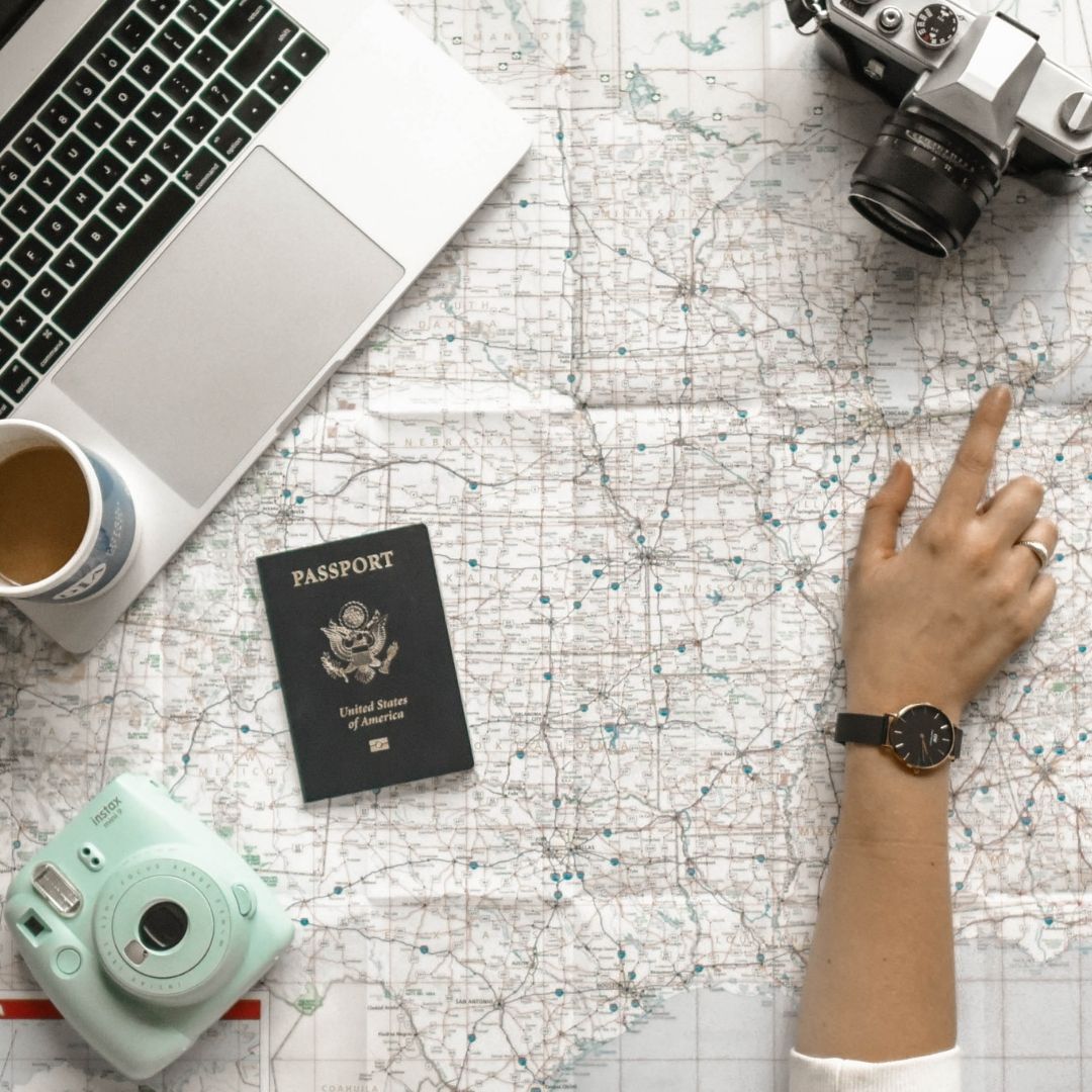A photo of a flat lay with a hand pointing to a large paper map of the United States. On top of the map are a Polaroid camera, USA passport, cup of tea, open laptop, and camera