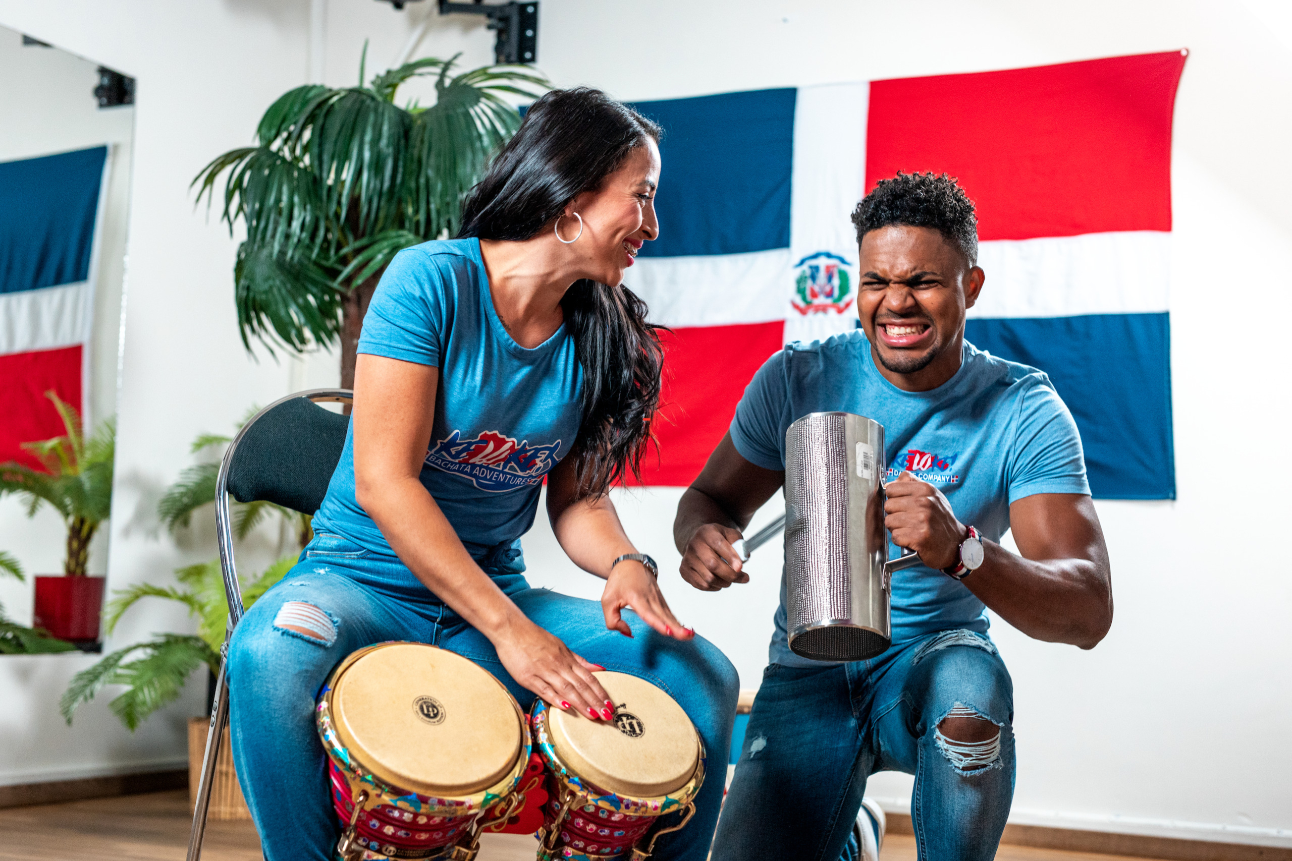 Junior and Carolina playing the bachata instruments, guira and bongo. The Dominican flag is in the backdrop.
