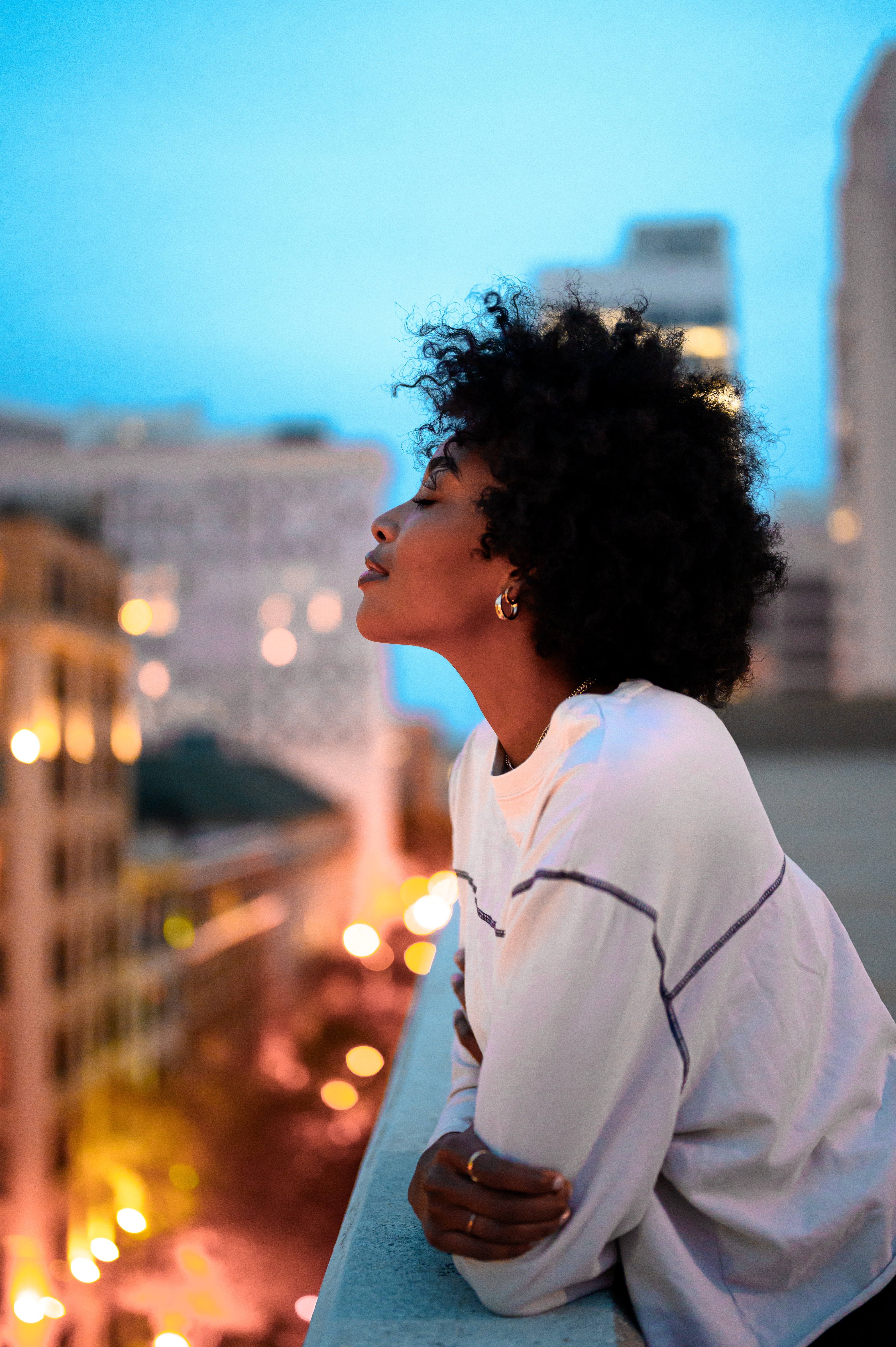 black woman with closed eyes leans against a balcony railing with evening city scene in background