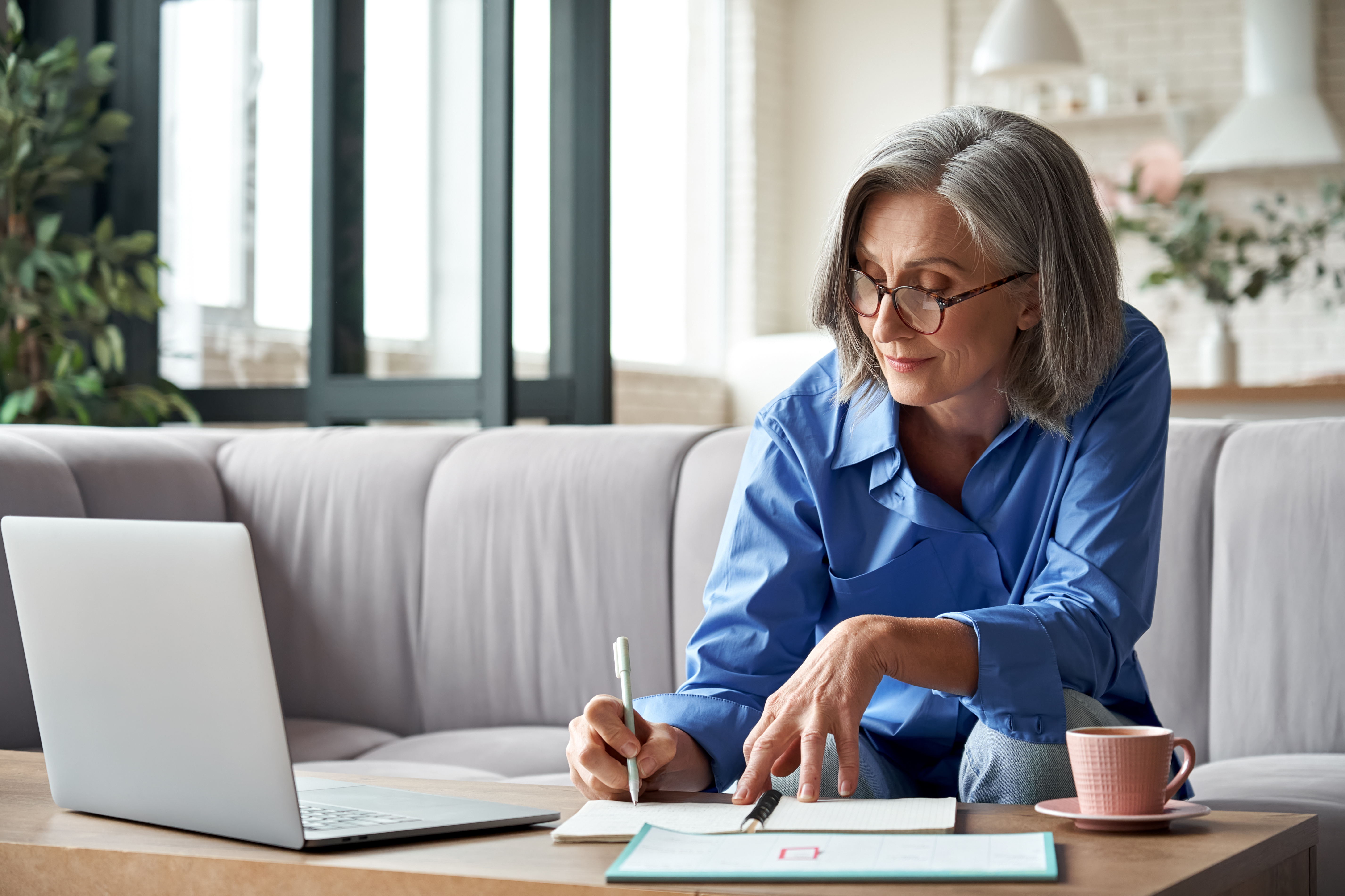 Middle aged woman takes notes, as she watches a video on a laptop.