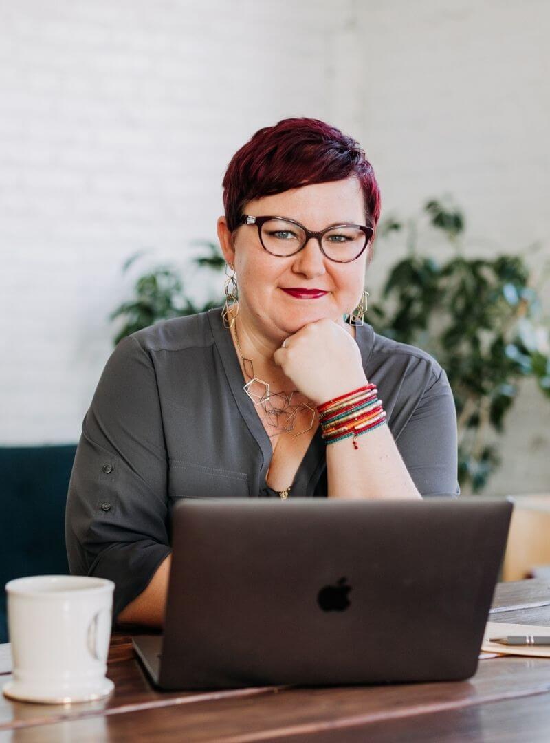 Cathy Hannabach wearing a grey shirt, glasses, and multicolored bracelets, resting her chin on her hand in front of a laptop
