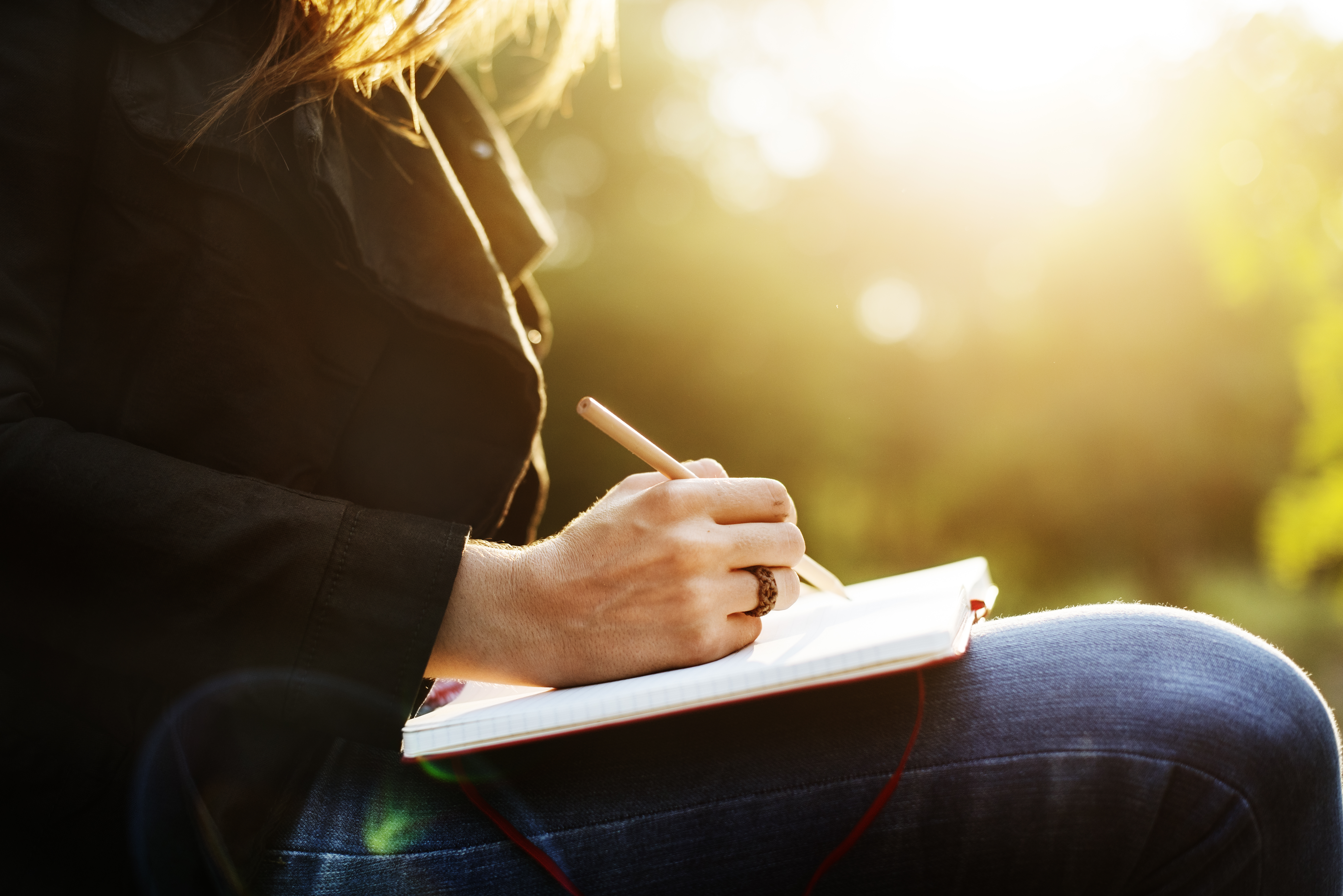 Photo of a woman writing in a notebook on her lap.  She is outside.