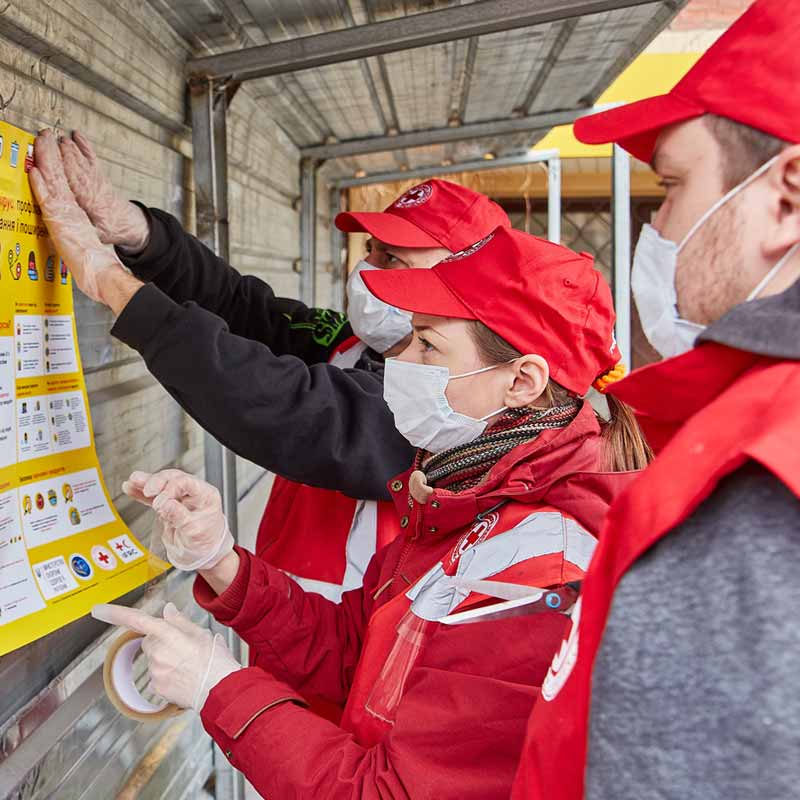 A group of people are putting up a yellow poster promoting safety information from the Canadian Red Cross. The people are wearing face masks, safety gloves, and Canadian Red Cross caps and jackets.
