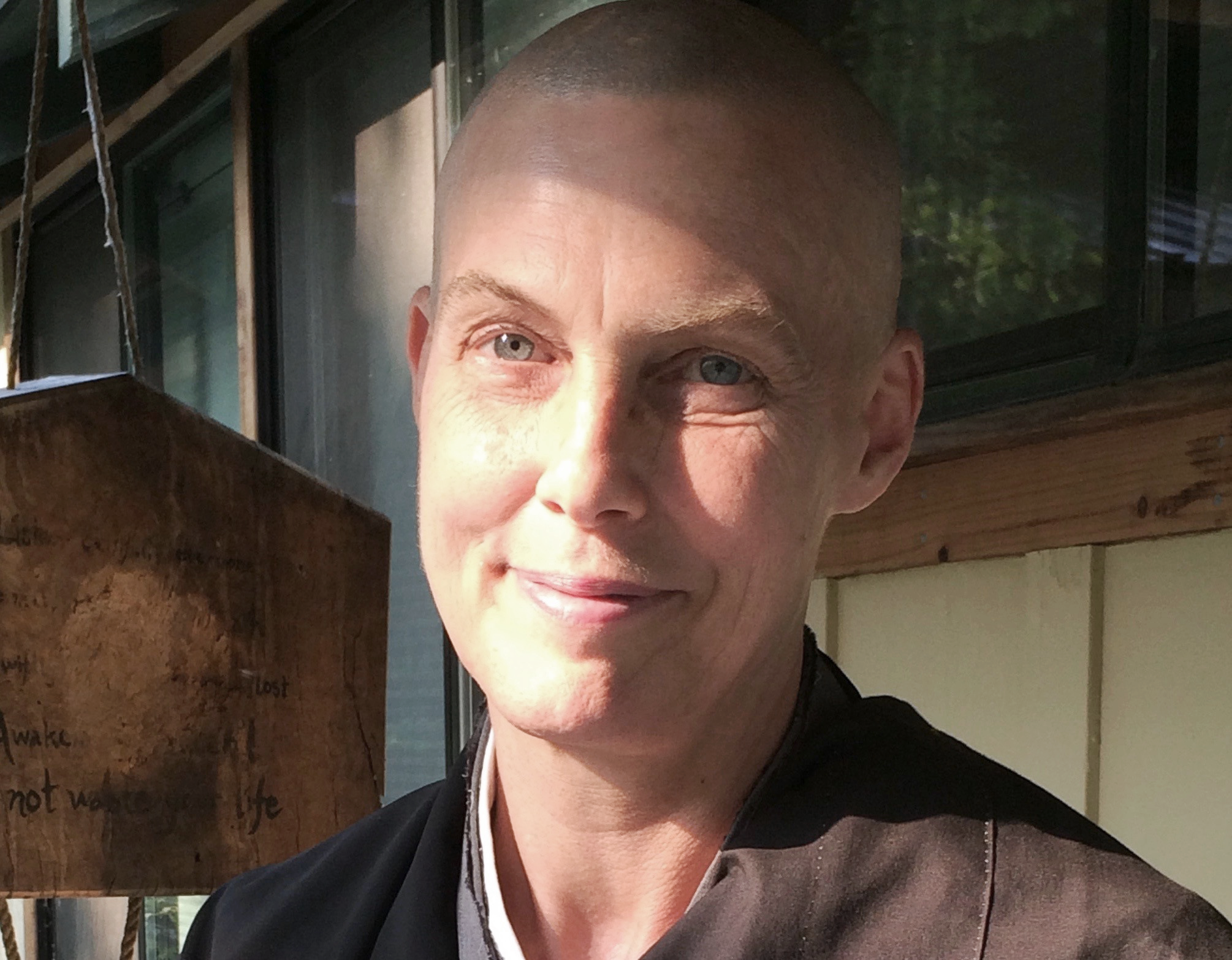 The image shows a smiling person with a shaved head and fair skin, wearing a dark robe, indicative of a Zen priest, in a sunlit natural setting.