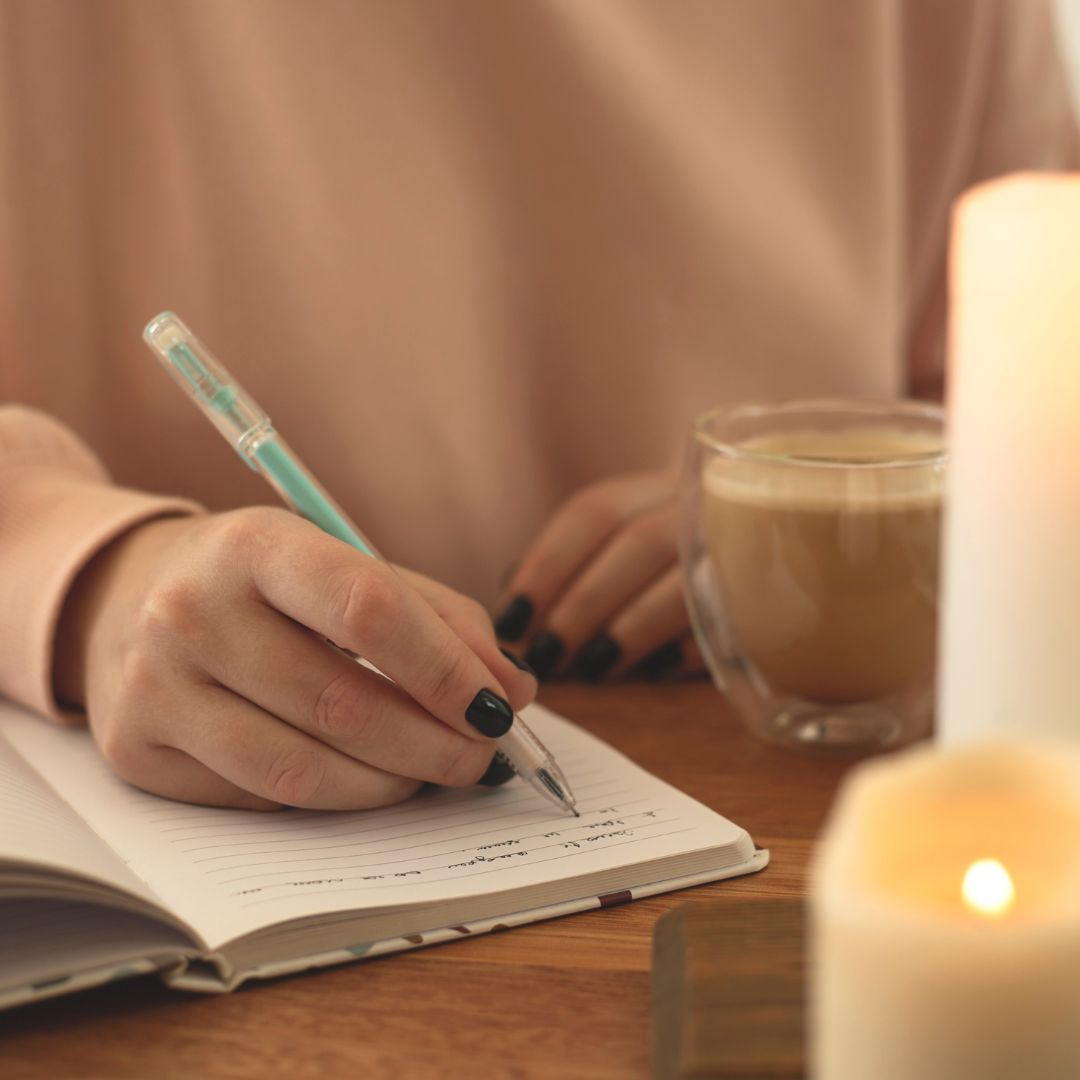 A woman writing in a journal. She has a cup of coffee and candles also in front of her.