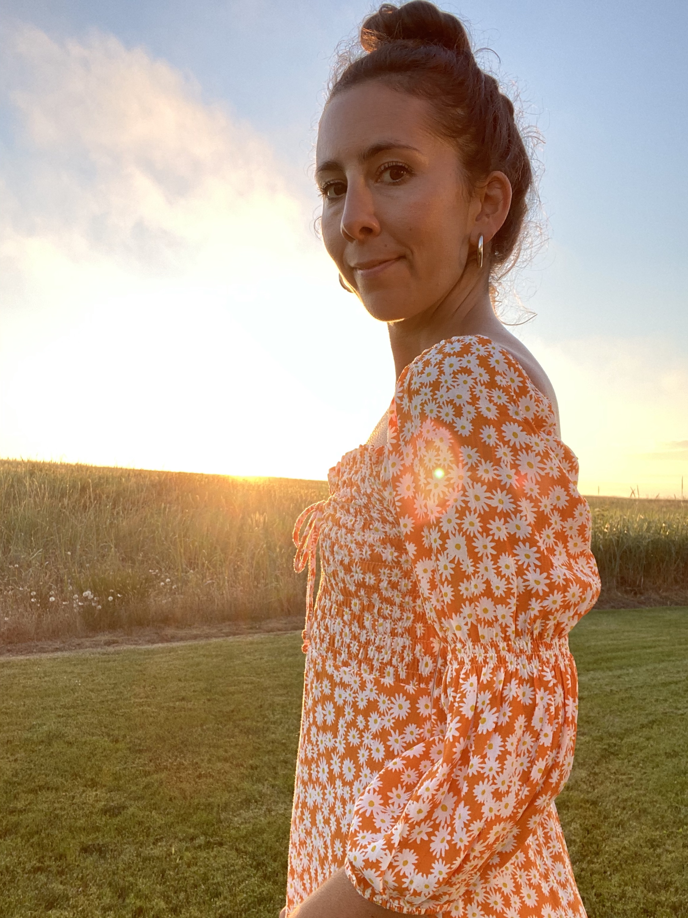 A woman standing in a field of grass feeling blissfully happy and in her worth. Wearing a orange daisy dress this is sustainably made.