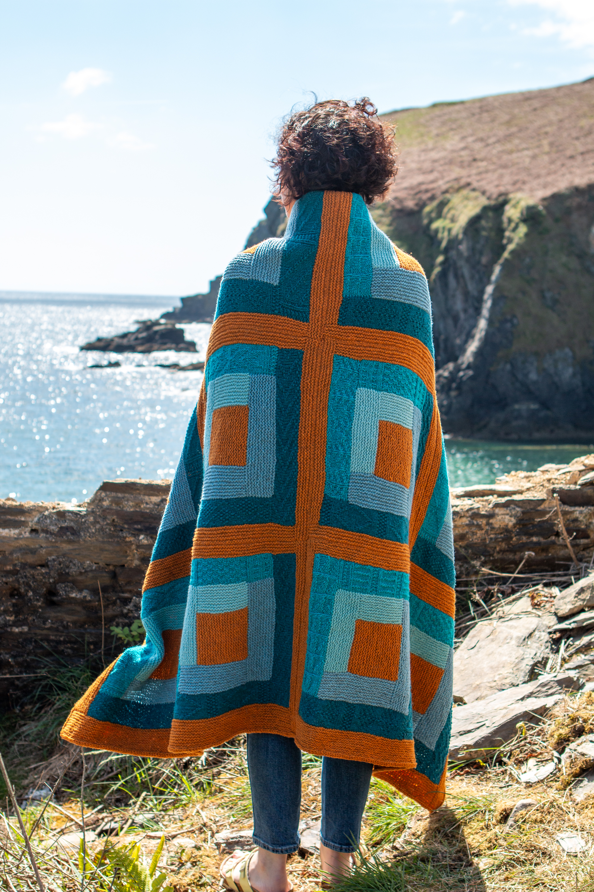 Woman with blanket thrown around her shoulders, standing on a cliff looking out to sea. 