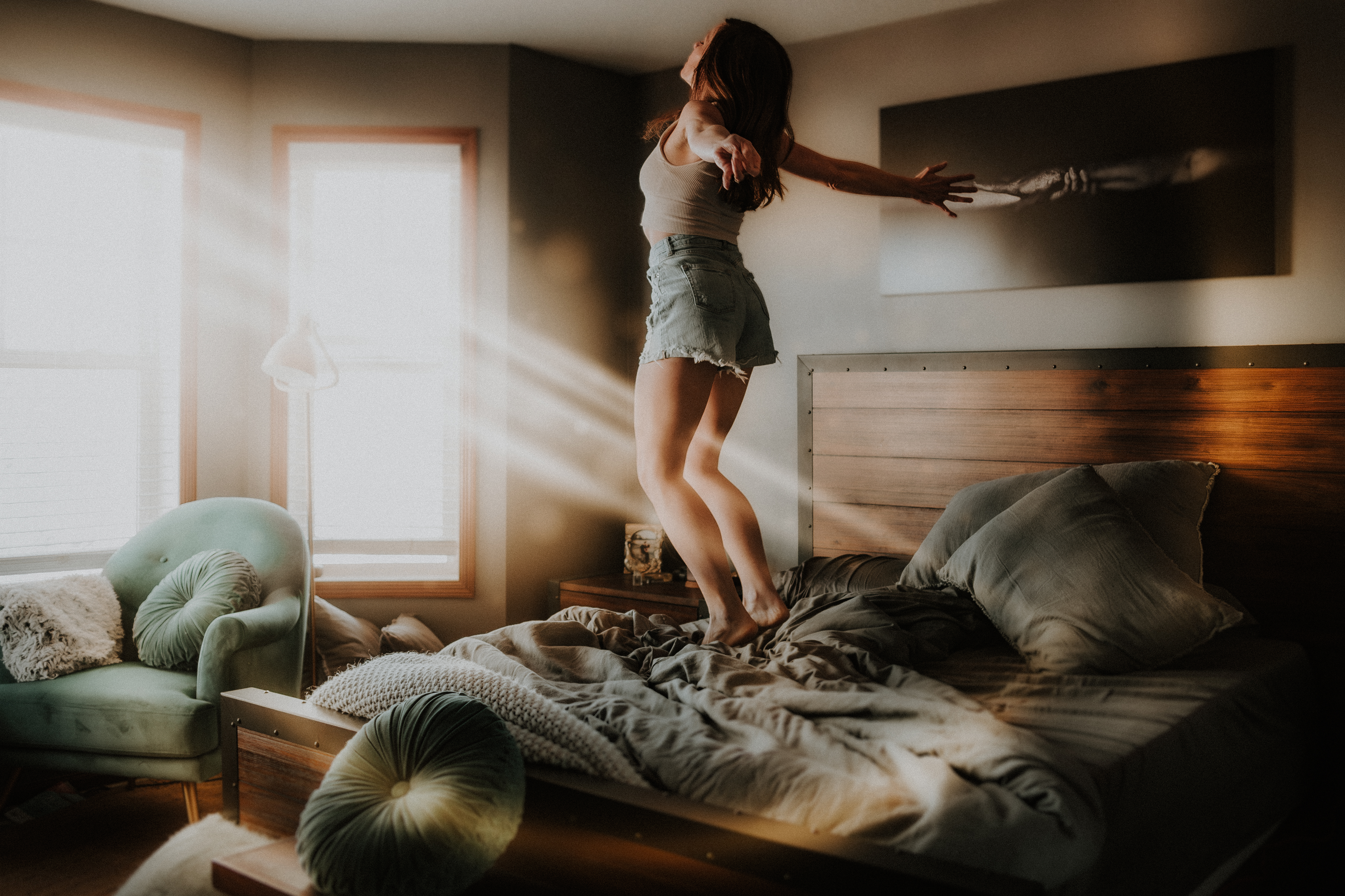 Self-portrait of a woman jumping on her bed towards beautiful light streaming in, showing resilience. This self-portrait photography course is Deaf and Hard of Hearing friendly