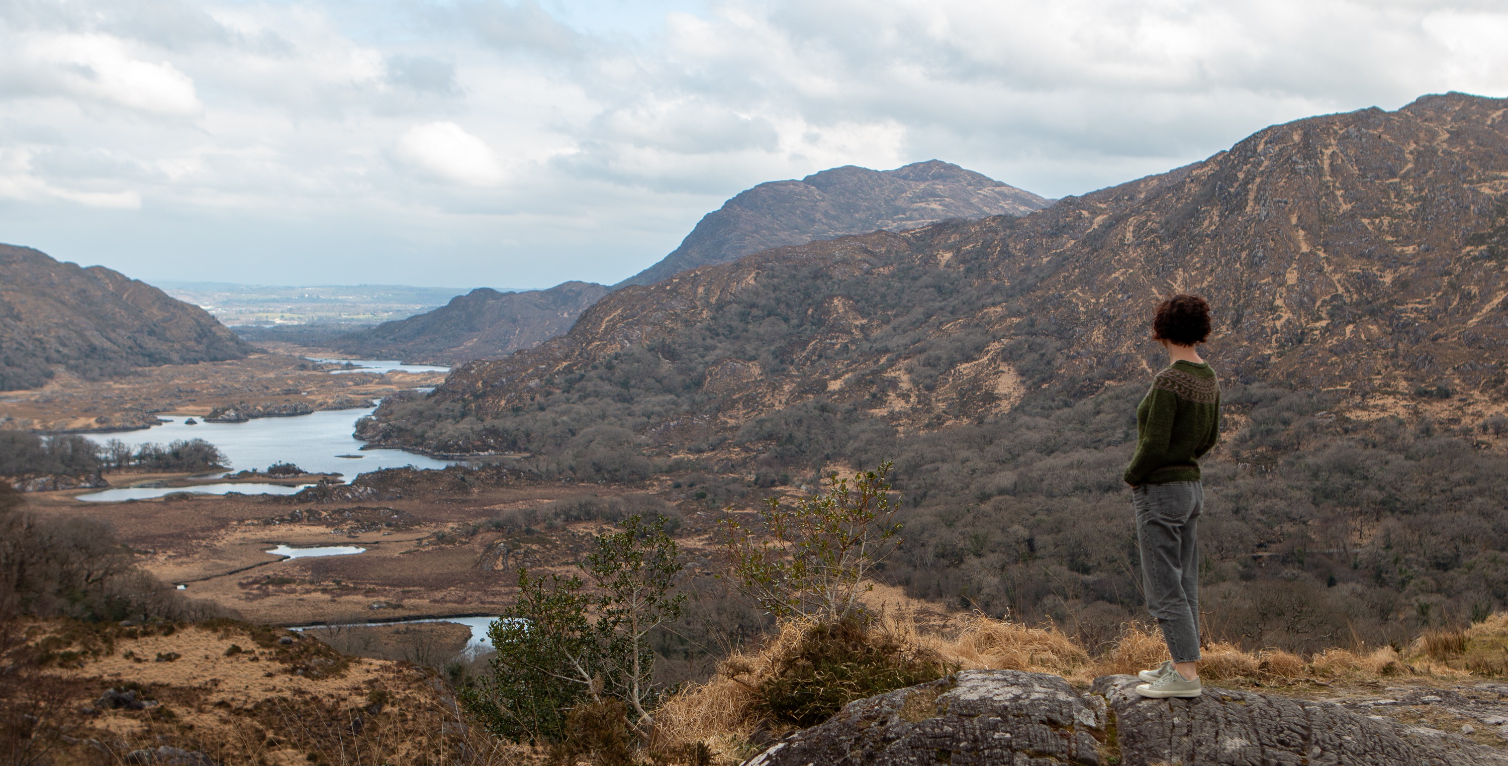 Carol Feller wearing the Daingean Cardigan, while stood on a rock looking out over a lake. 