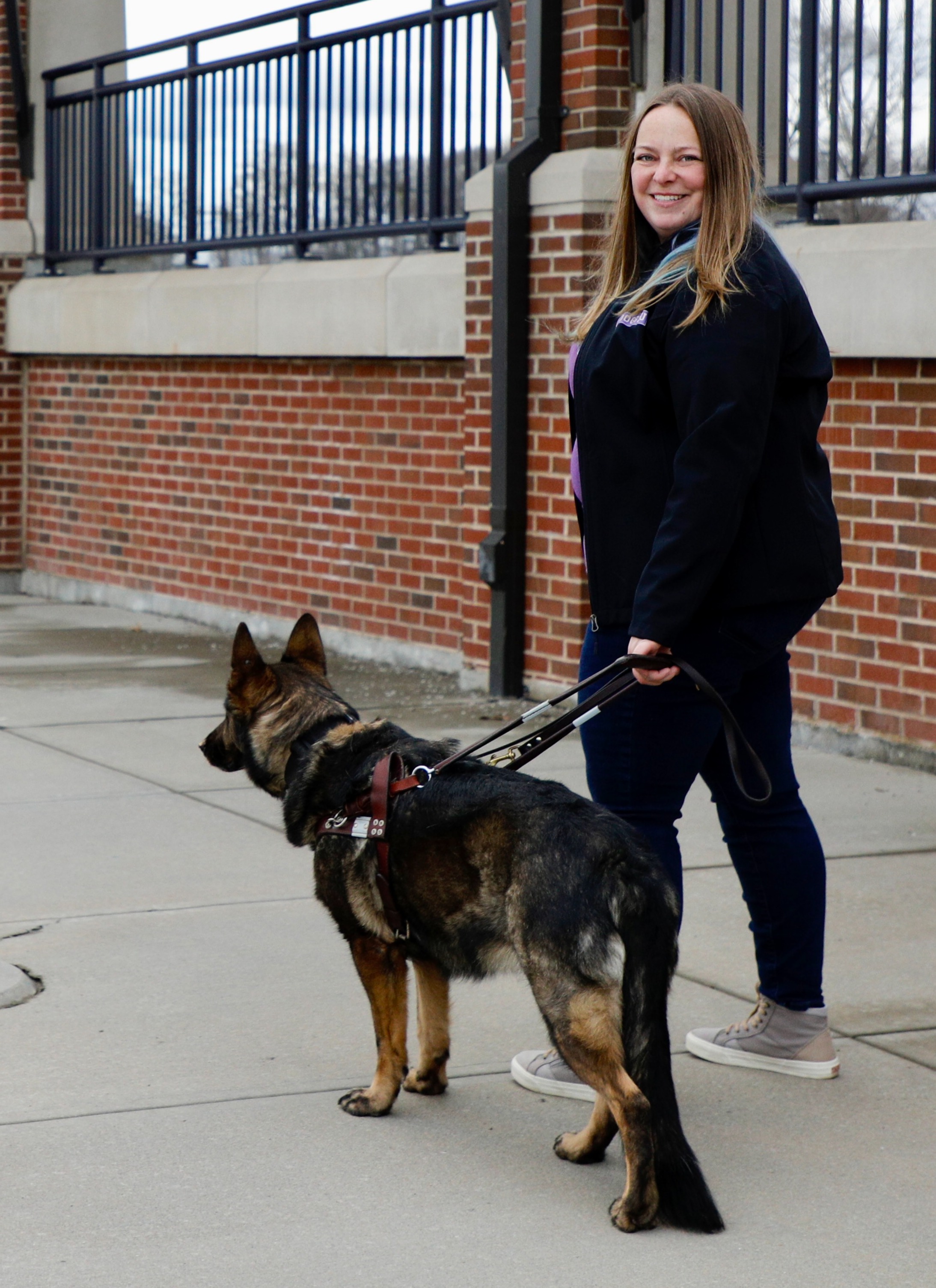 Light-skinned woman with blonde and blue hair looks back at camera wearing jeans and a black coat. She holds the handle of a german shepherds guide harness in her left hand