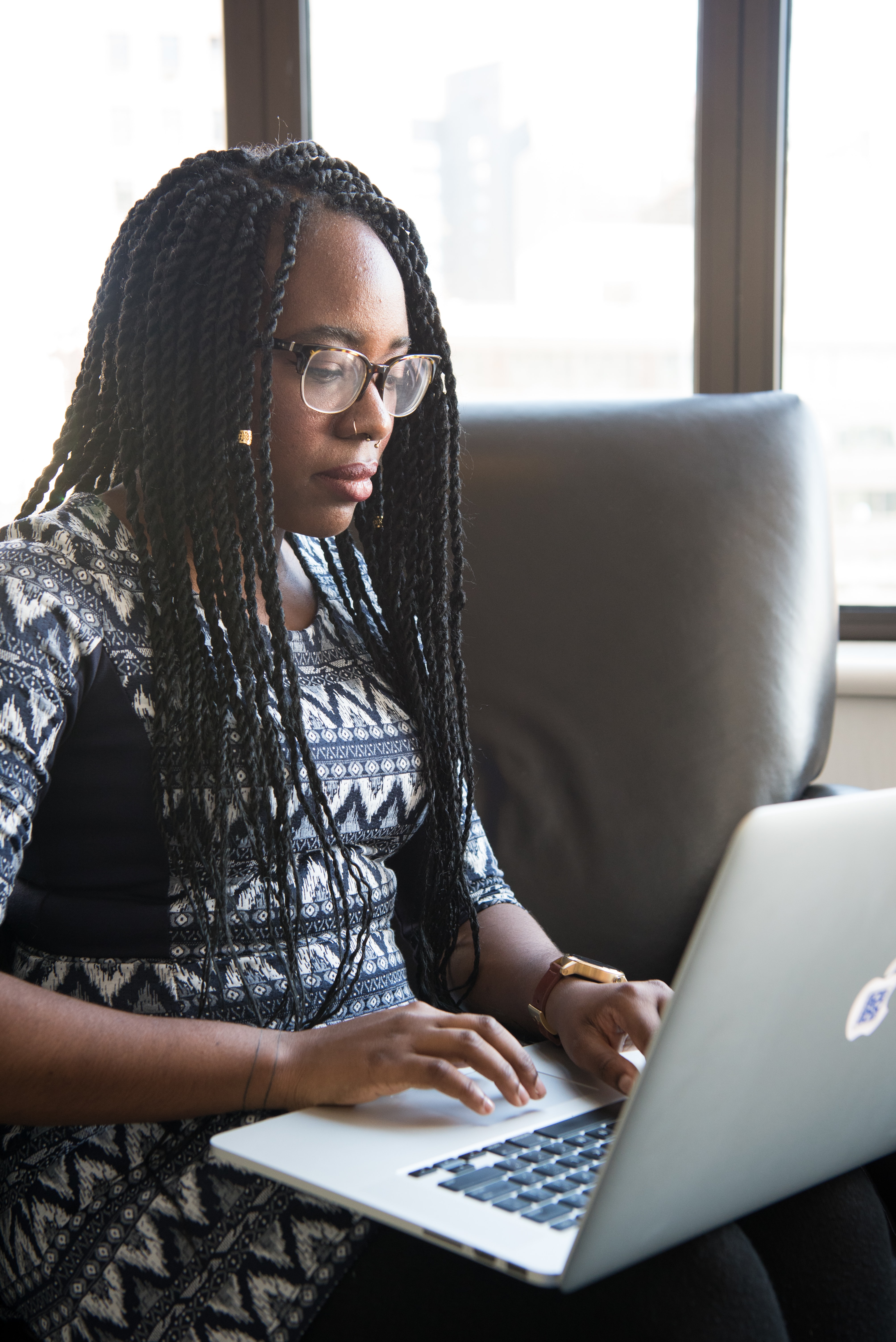 Black woman with glasses wearing a patterned dress on a chair with an open laptop on her lap