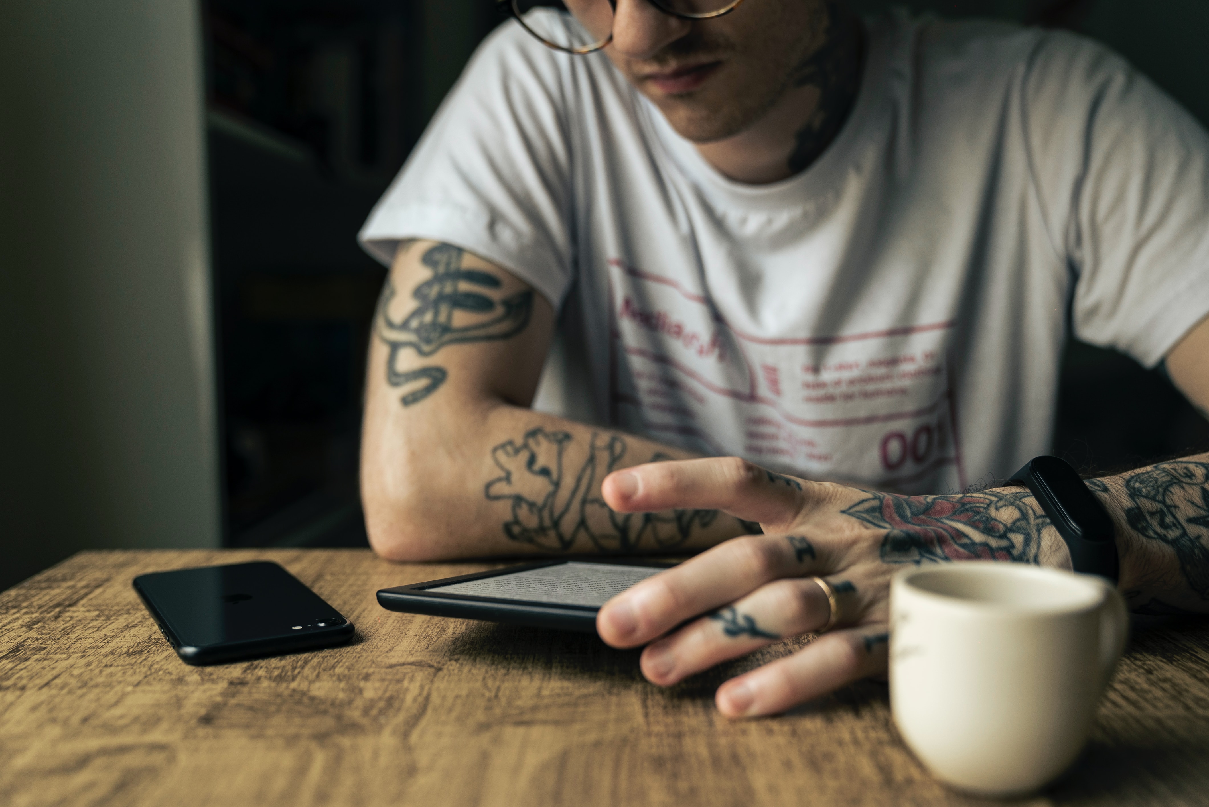 Background is of a white male with glasses and a white shirt, reading on his kindle with a cup of coffee on his left, and his phone on his right. He is sitting at a wooden table, and he has tattoos up both arms and hands.