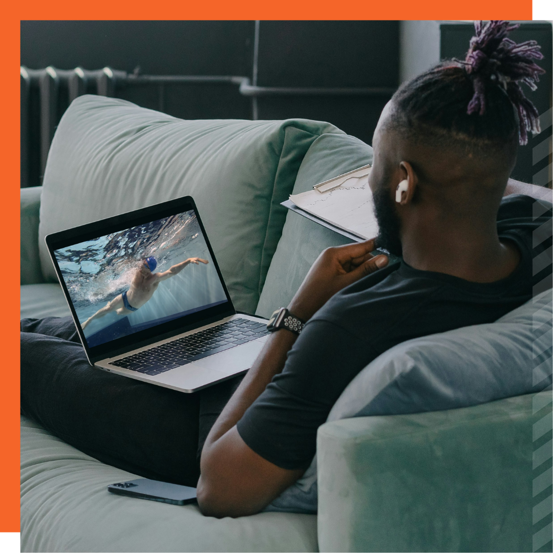 Image of a man sitting on a sofa watching a swim drill video on a laptop.