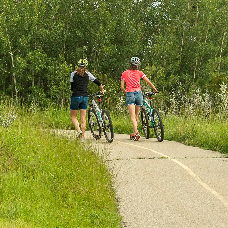 Two people wearing helmets are walking their bikes on a sunny summer day.