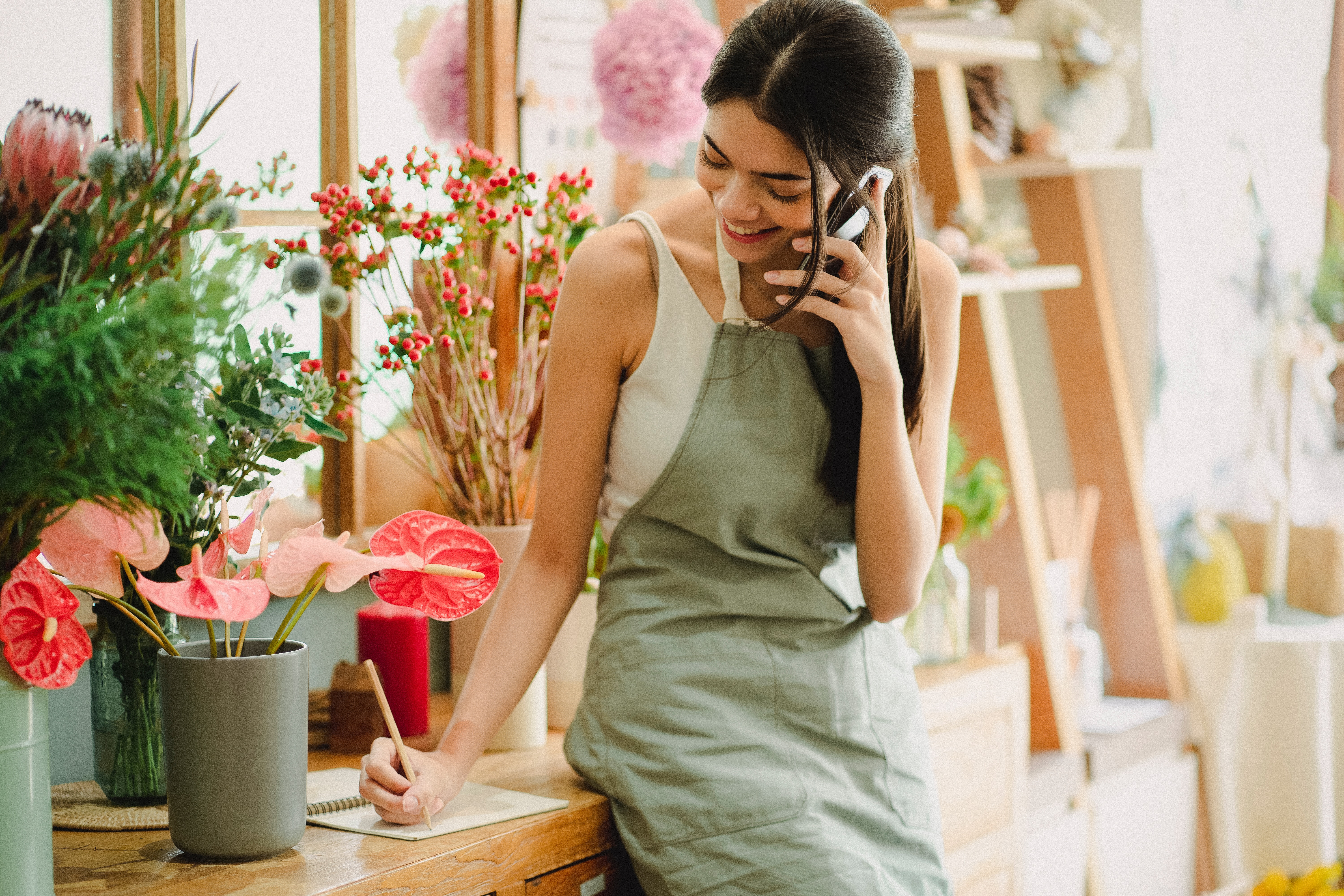Photo of a young woman on the phone and writing on a note pad. She is wearing an olive apron and works in a flower shop.