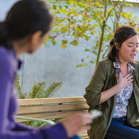 Person sitting on a bench holding their chest and struggling to breathe while another person approaches.