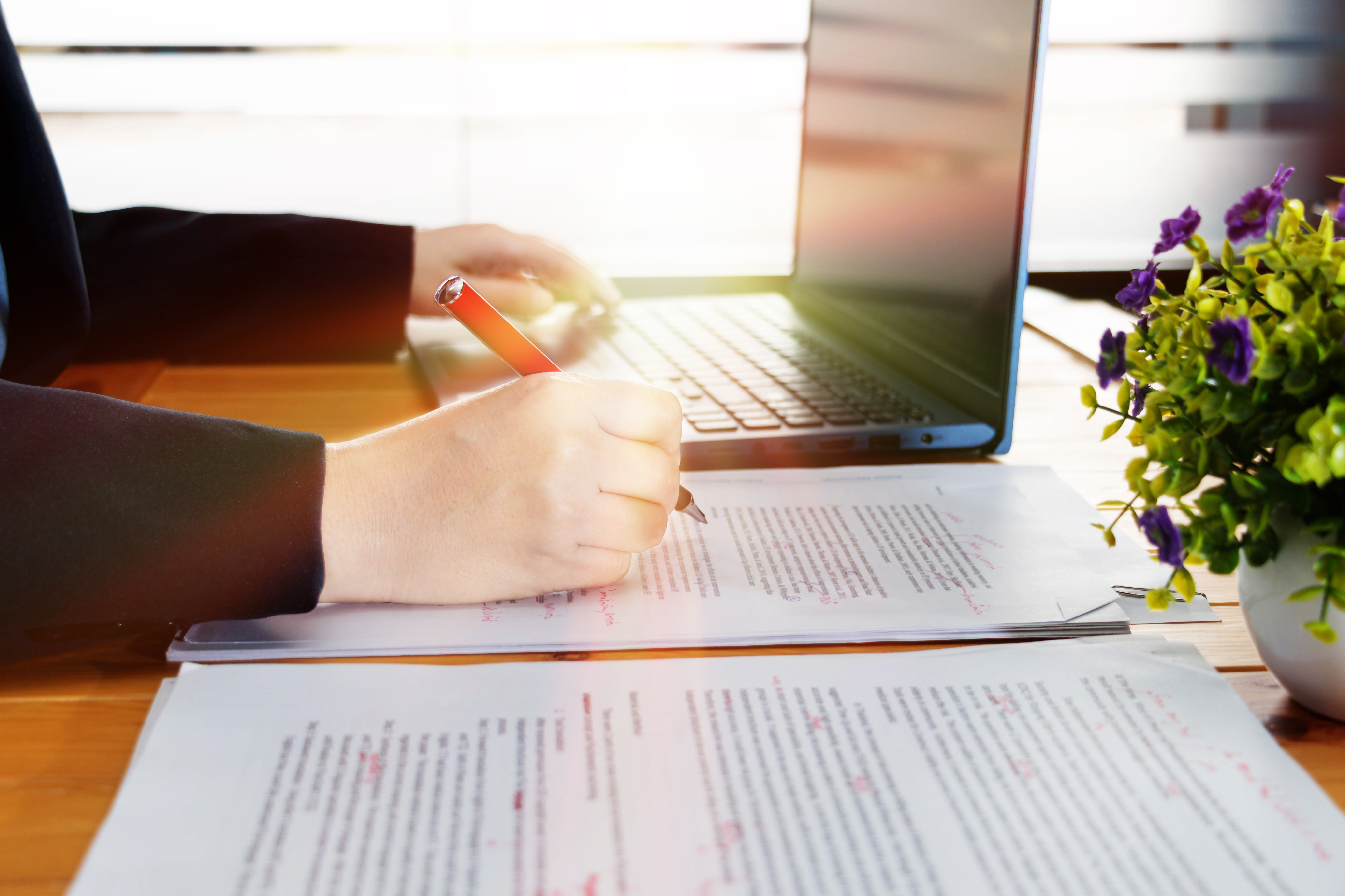 Woman with a laptop holding a red pen and marking up a paper document, with a small vase of flowers on her desk