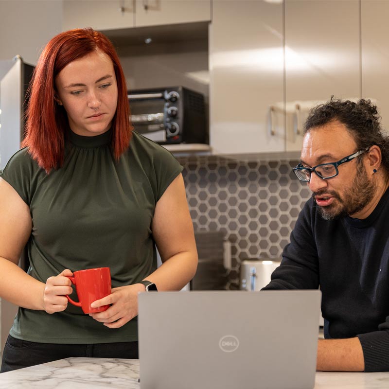Two people looking at a laptop, one is showing the other some important information on the computer screen. They both look thoughtful and are in the kitchen.