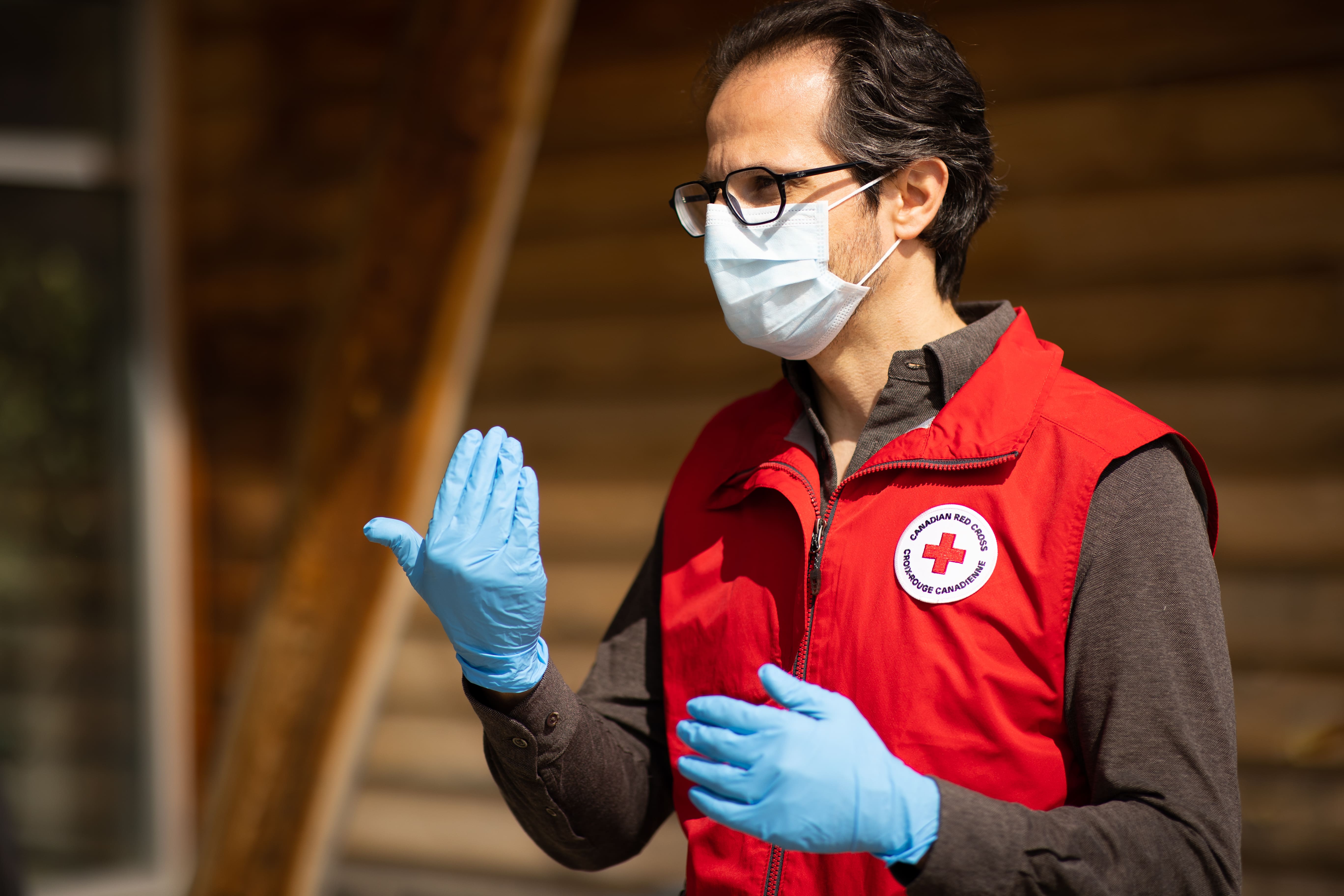 A volunteer wearing a Canadian Red Cross vest, facial mask, and gloves directs others to approach.