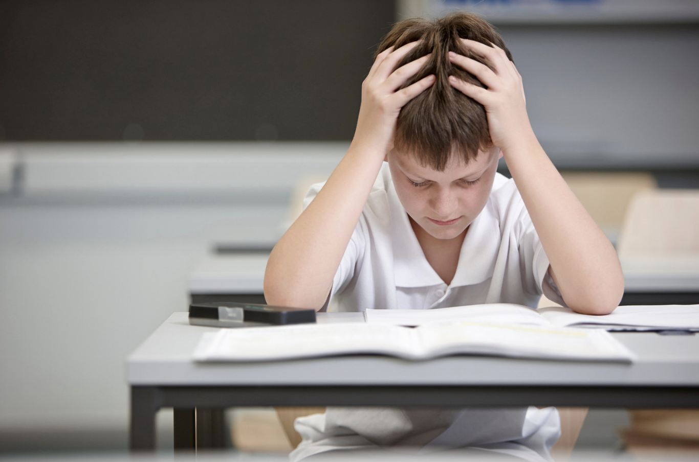 A young boy sits at a desk with his head in his hands.