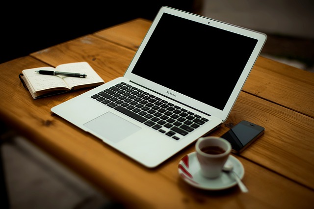 An open laptop computer, an open blank book with a pen, and a cup of coffee. 