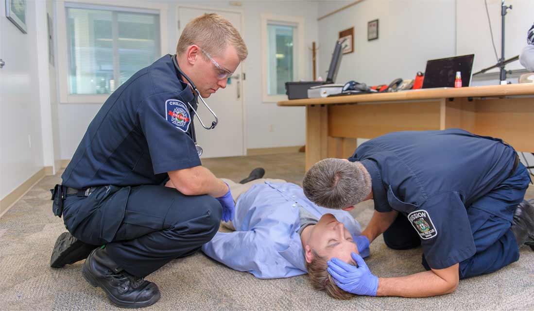 A person wearing office clothes lying in the floor possibly suffering from opioid poisoning due to pale face, while two paramedics are assisting it. One paramedic is checking for breathing and the other paramedic is kneeling at the side. The place appears to be an office workplace.