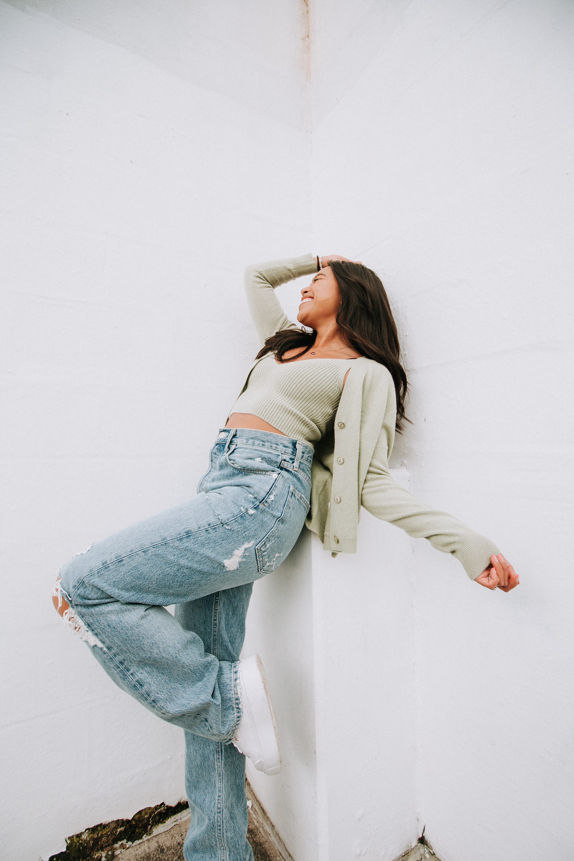 Leaning against a white wall, wearing 90s jeans and a matching green top and cardigan