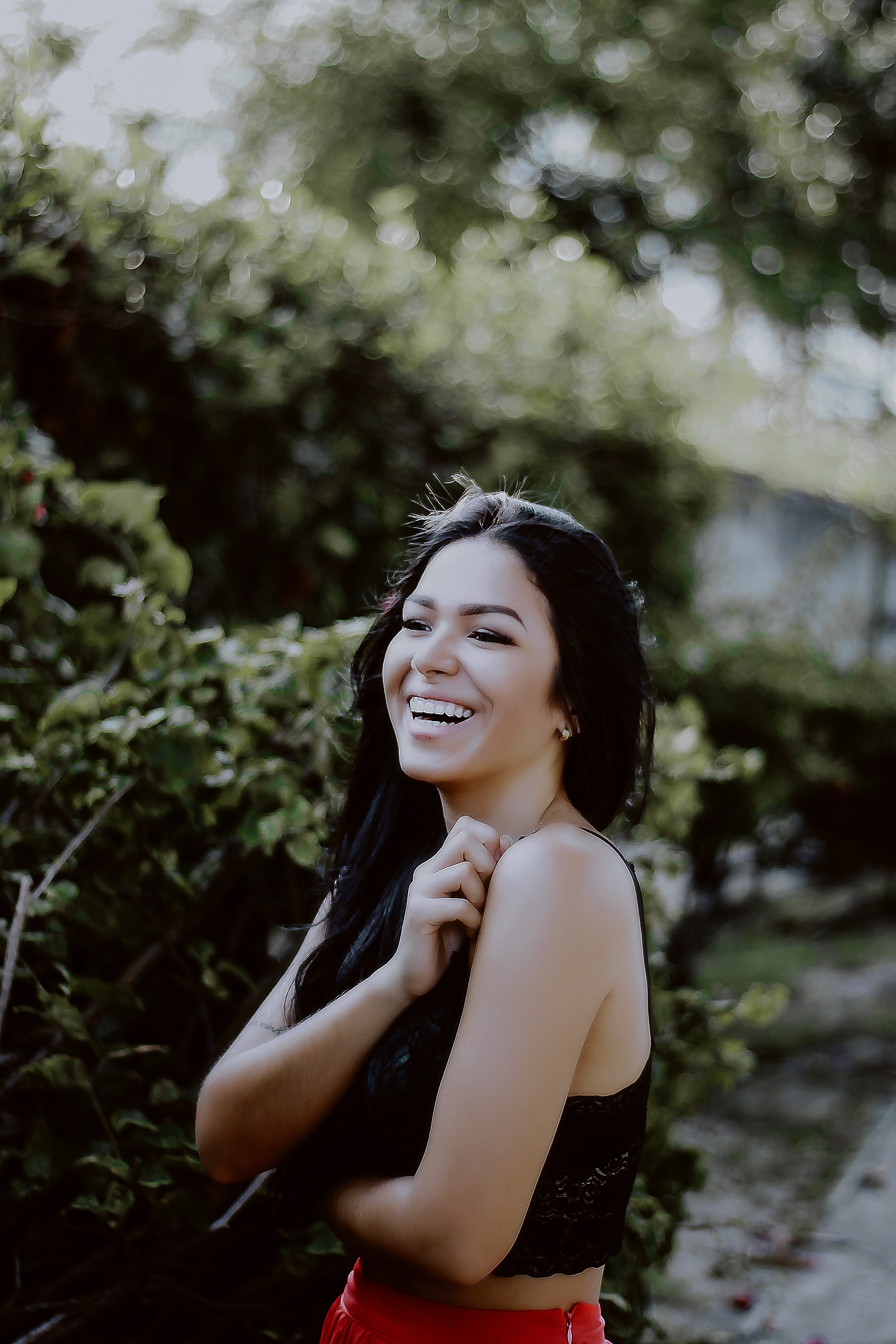 person with long dark hair hugging herself, smiling, wearing black tank top and red skirt, in front of shrubs and trees