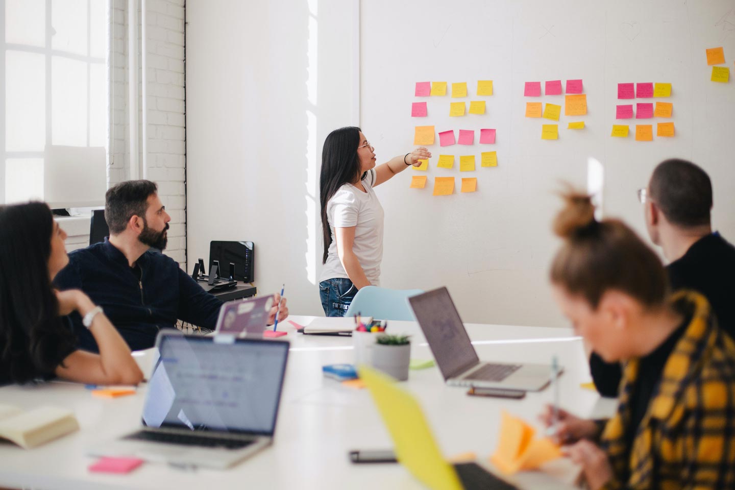 A woman places sticky notes on a wall while in a meeting with four other people.