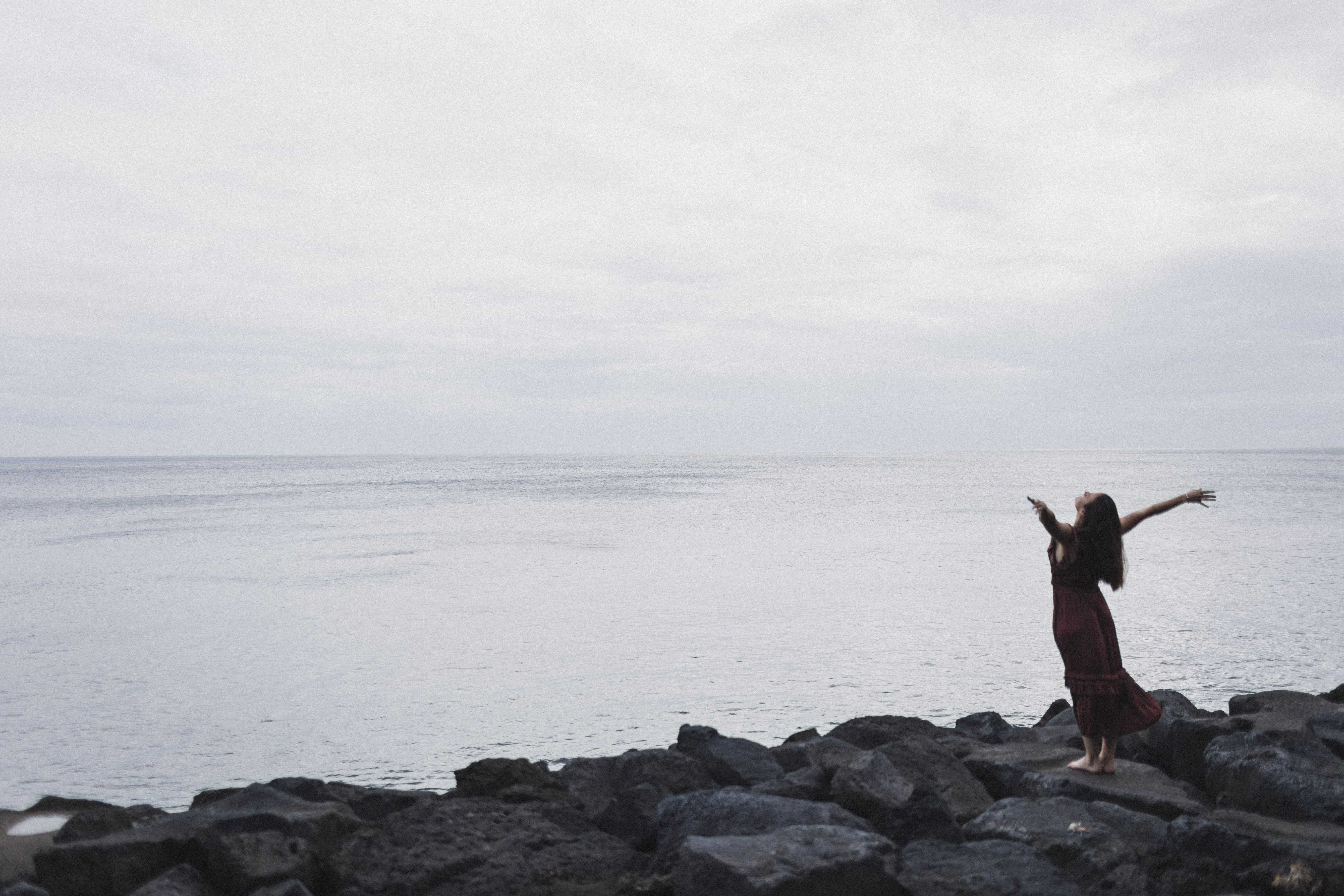 woman standing on volcanic rocks, arms outstretched towards the ocean