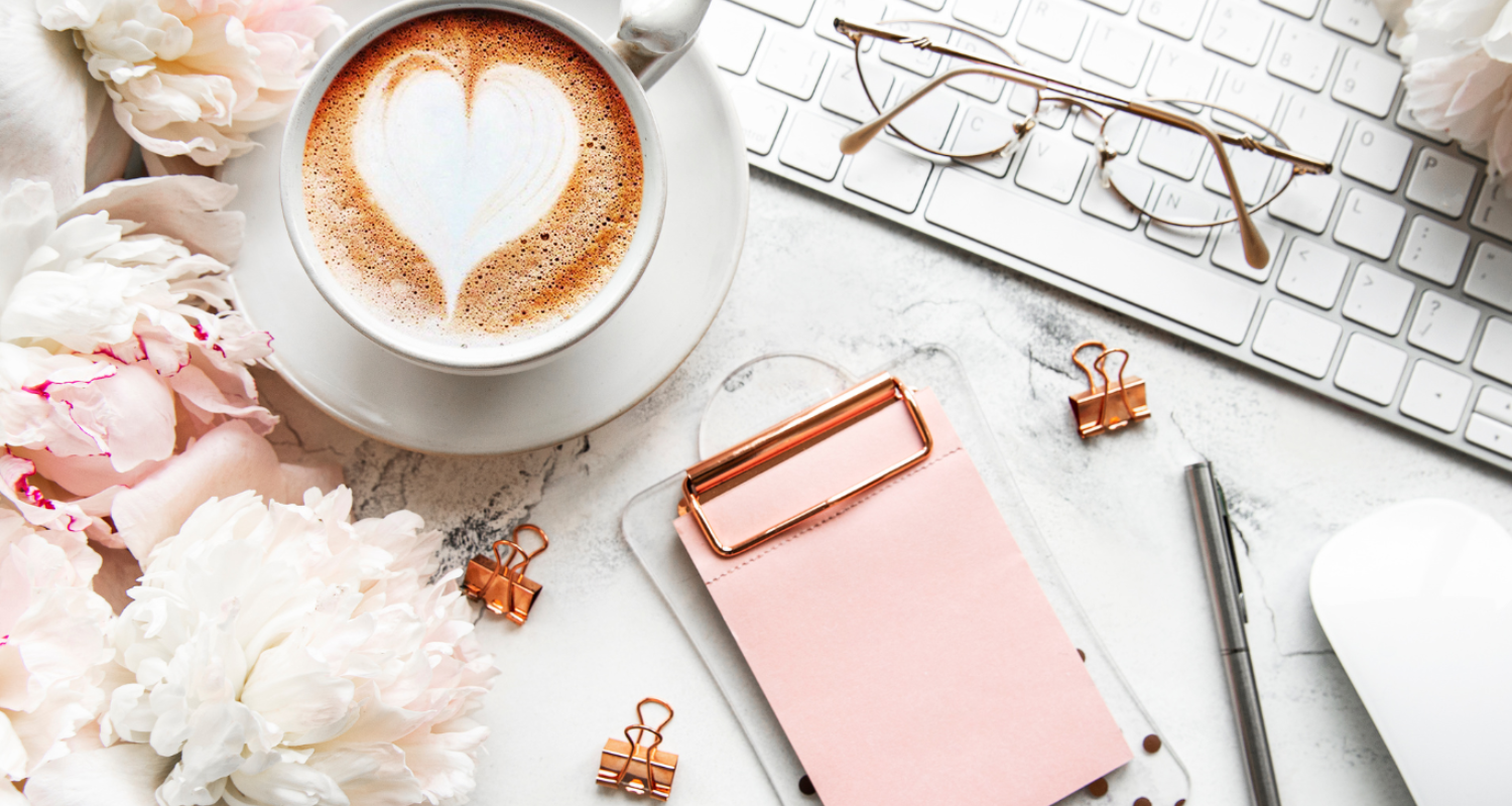 Coffee cup with a heart stenciled in froth, on a desk with a keyboard, glasses and small clipboard