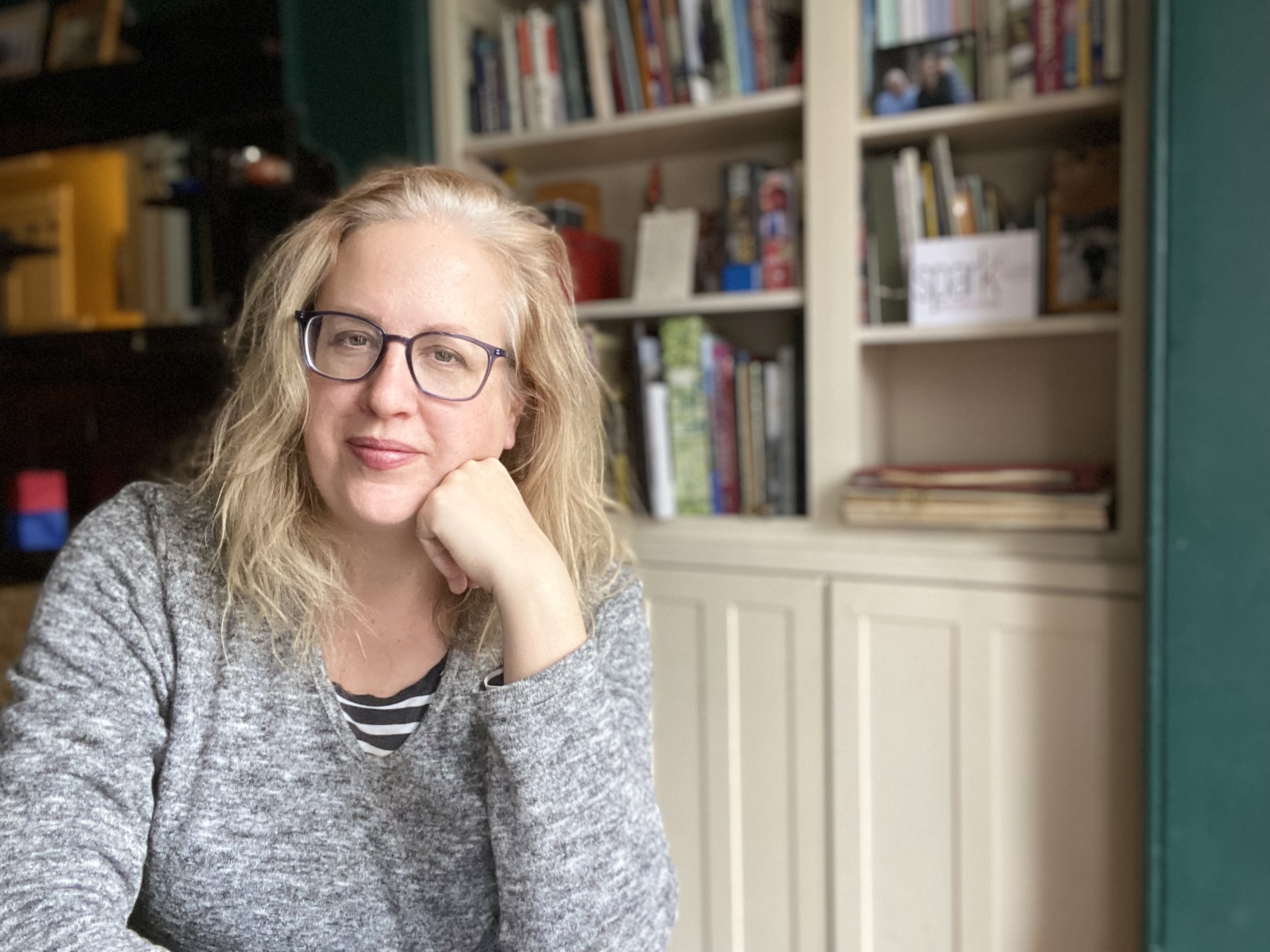 A blond woman wearing glasses is smiling, her chin leaning on her hand, with a wall of bookshelves behind her.