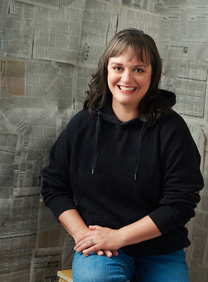 A woman wearing blue jeans and a black pullover hoodie sits on a wooden stool in front of a giant newspaper collage. She looks friendly and approachable as she smiles right at the viewer.