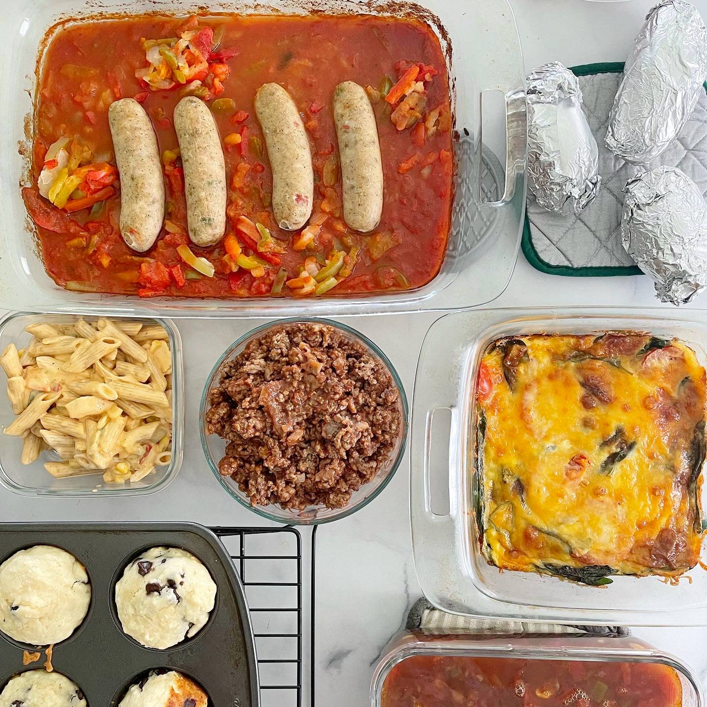marble surface with variety of food dishes (sausages on top of red sauce with veggies in rectangle dish on top left, baked potatoes in aluminum foil on silver and green hot pad in top right, dish of pasta in bottom left, circle dish of ground meat in bottom middle, and cheese-topped veggie egg bake in square pan in bottom right corner)